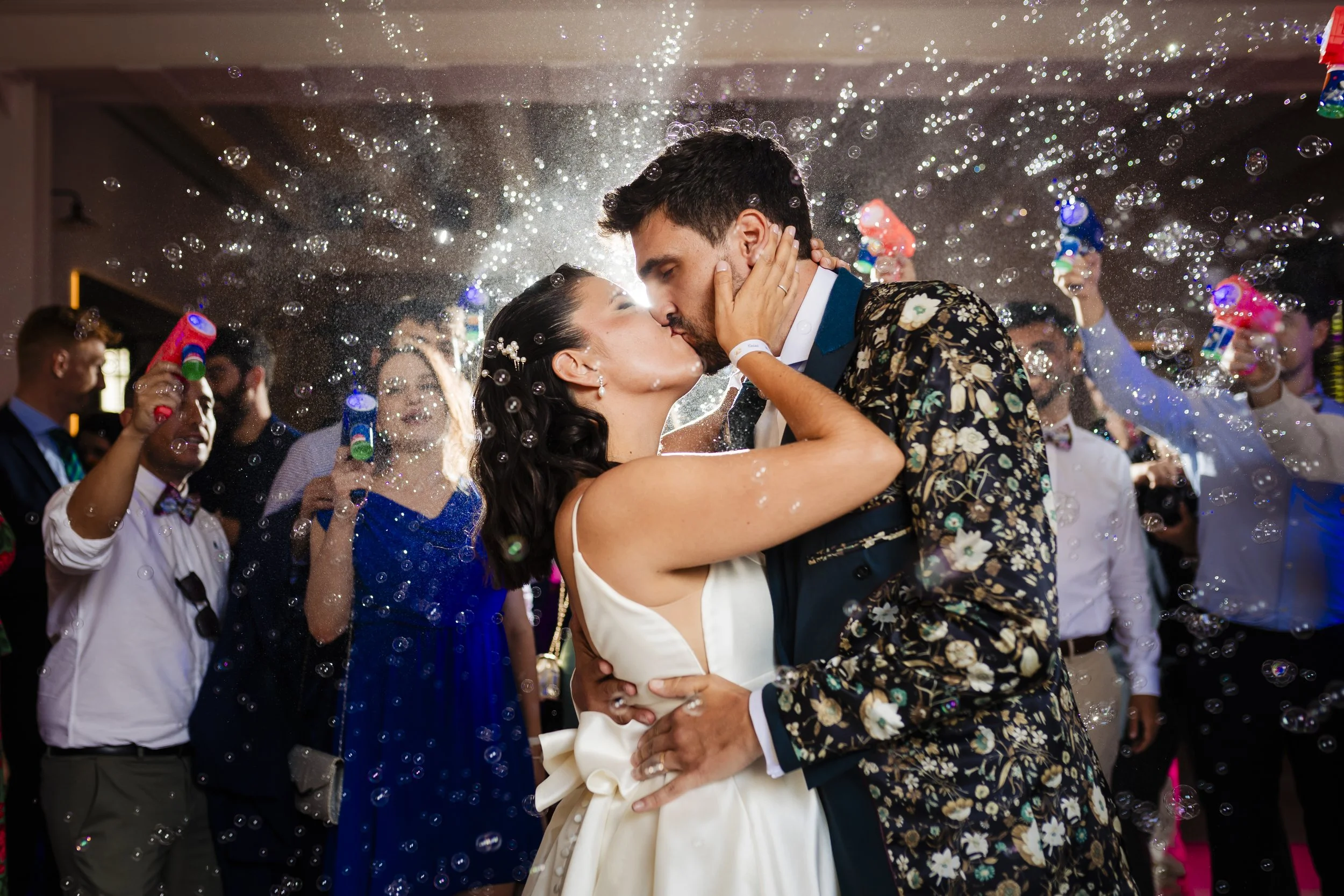Una pareja besándose en su boda, rodeados de invitados con aspersores de agua y burbujas.
