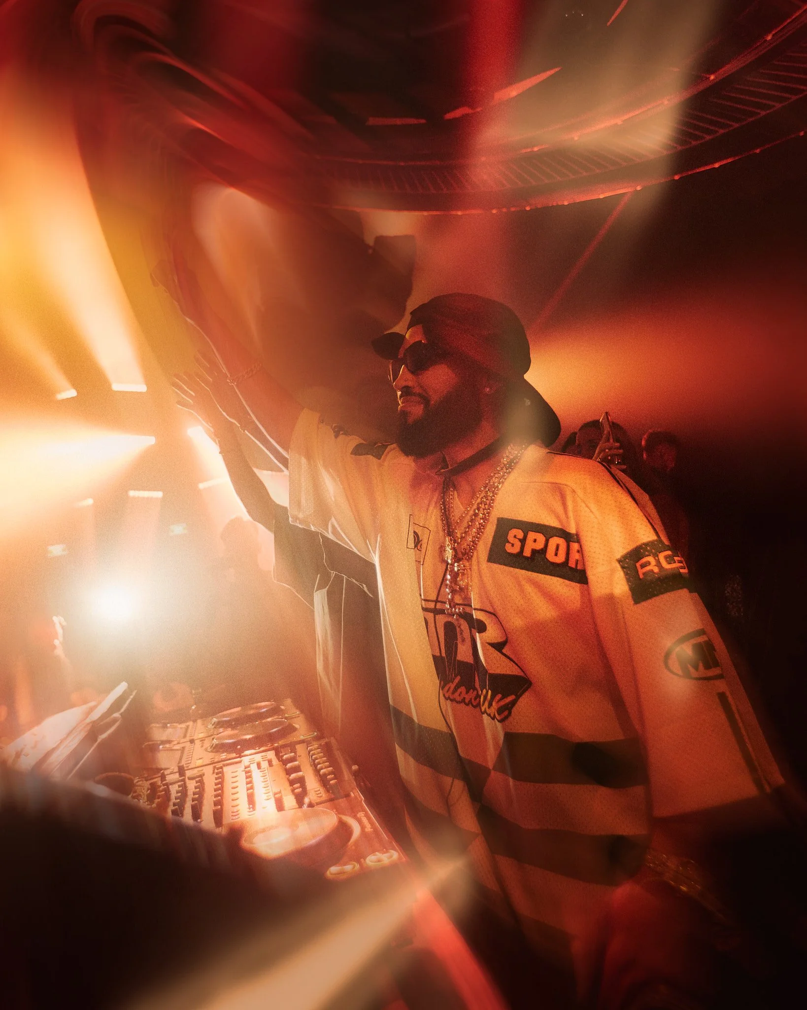Un DJ con gafas de sol, jerseys con varios logotipos y cadenas, tocando en una cabina de DJ en una fiesta con luces de colores.