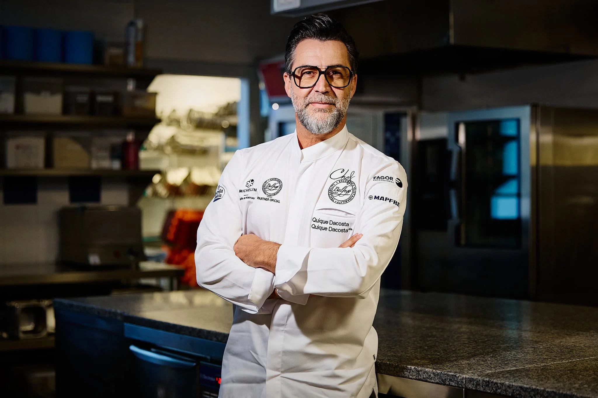 Un chef con barba y gafas, vestido con chaqueta blanca de chef, en una cocina, con los brazos cruzados