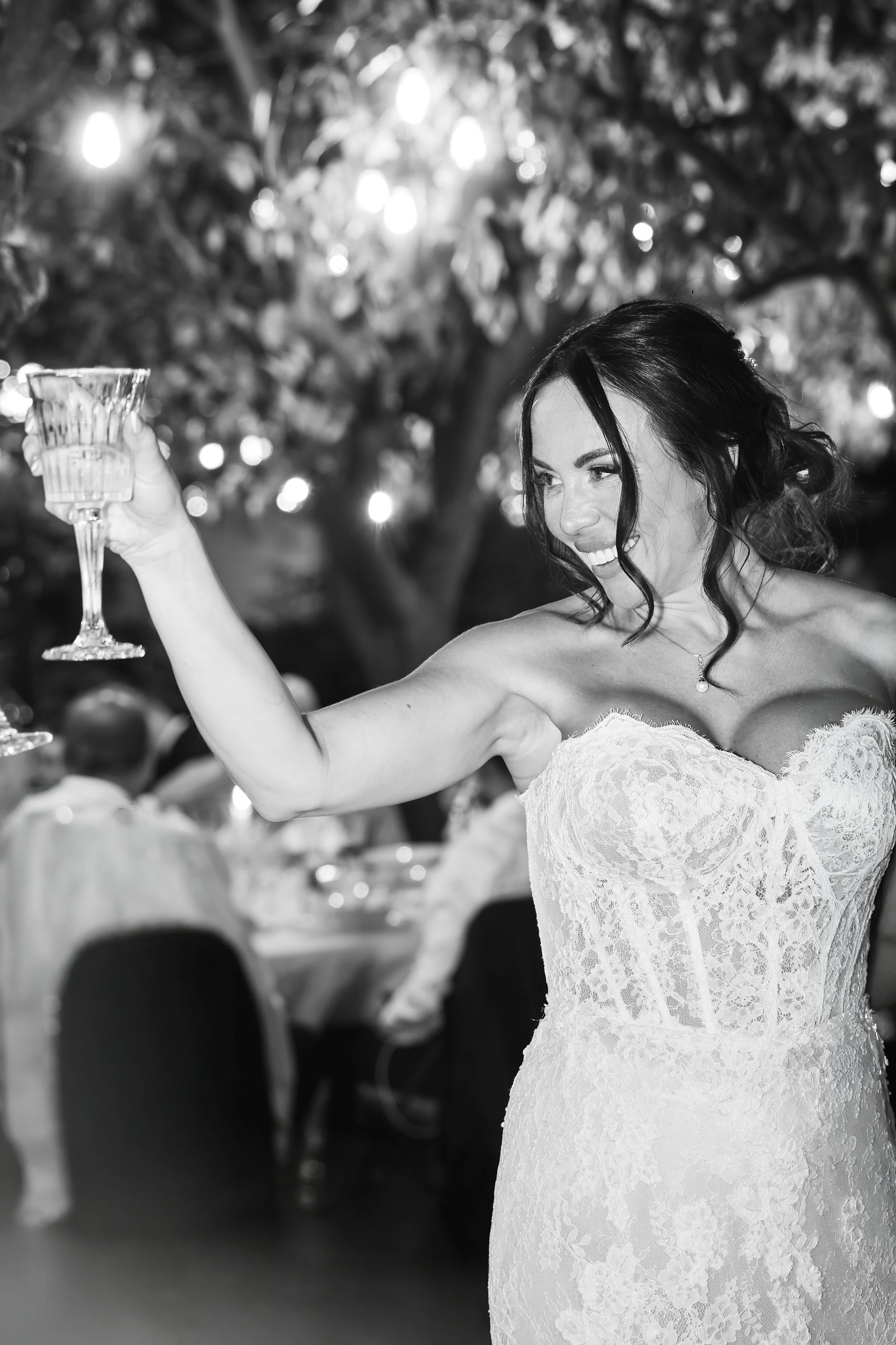 Mujer sonriendo en una boda, sosteniendo una copa en un evento con fondo de luces y árboles.