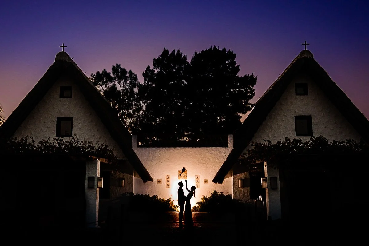 Pareja besándose al atardecer frente a edificios con cruces religiosos en el techo, con árboles de fondo.