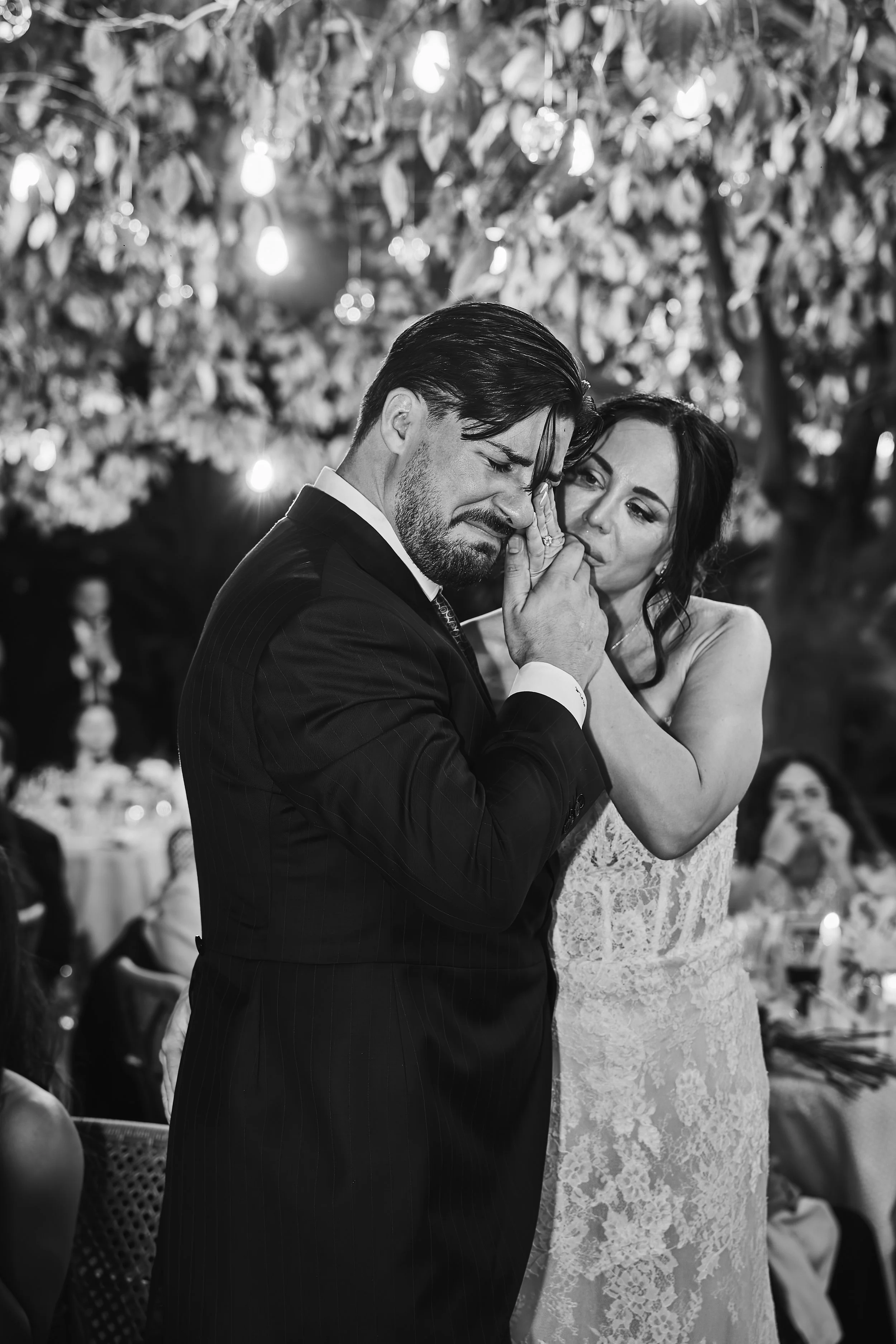 Pareja bailando en una boda, el hombre viste un traje negro y la mujer lleva un vestido de encaje, ambos en un momento emotivo, con decoraciones de luces y hojas en el fondo.
