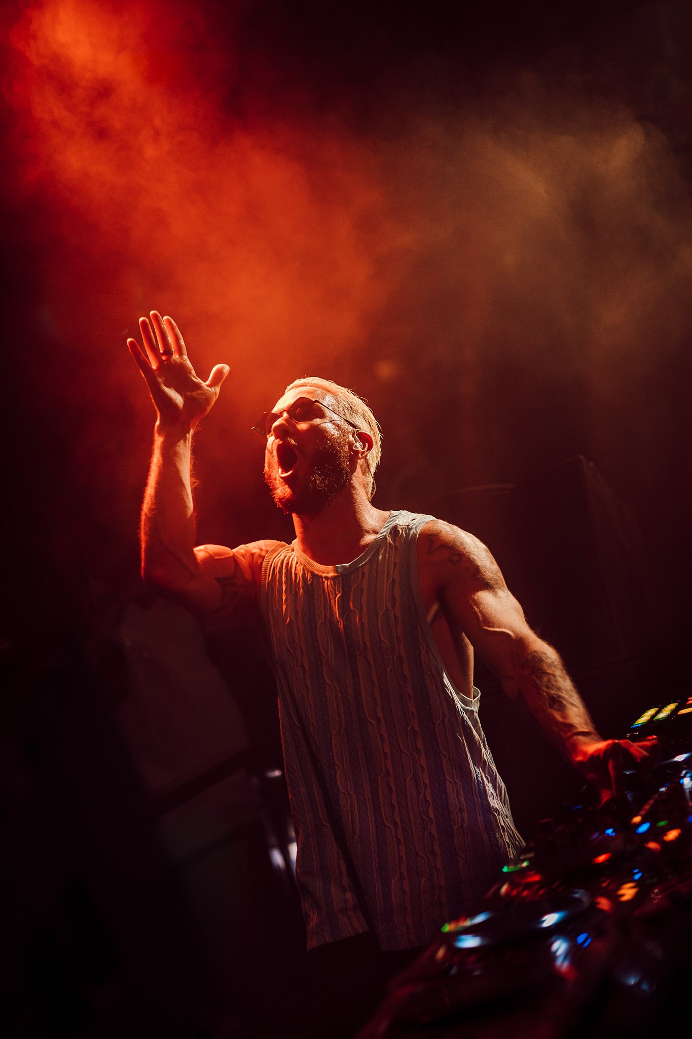 James Hype Un DJ con gafas y barba, con camiseta sin mangas, mezclando música en una fiesta con luces rojas y nubes de humo.