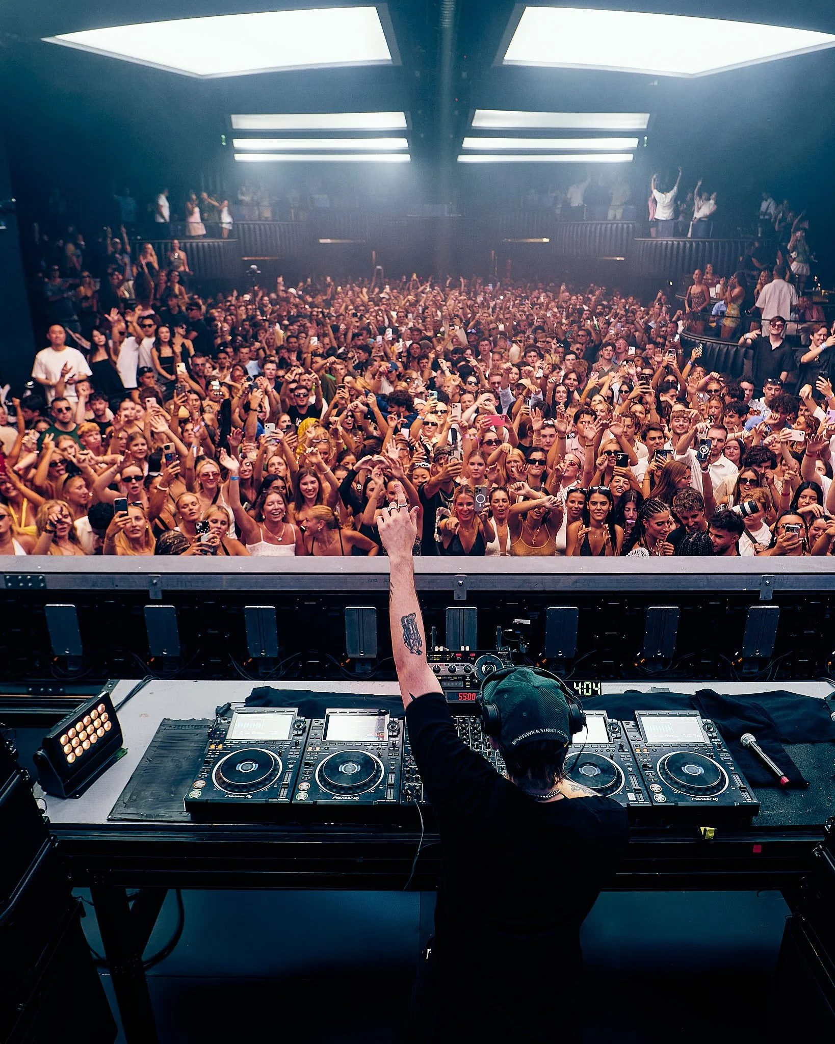 Un DJ en cabina frente a una multitud en un concierto, levantando una mano, en un entorno oscuro con luces brillantes y humo.
