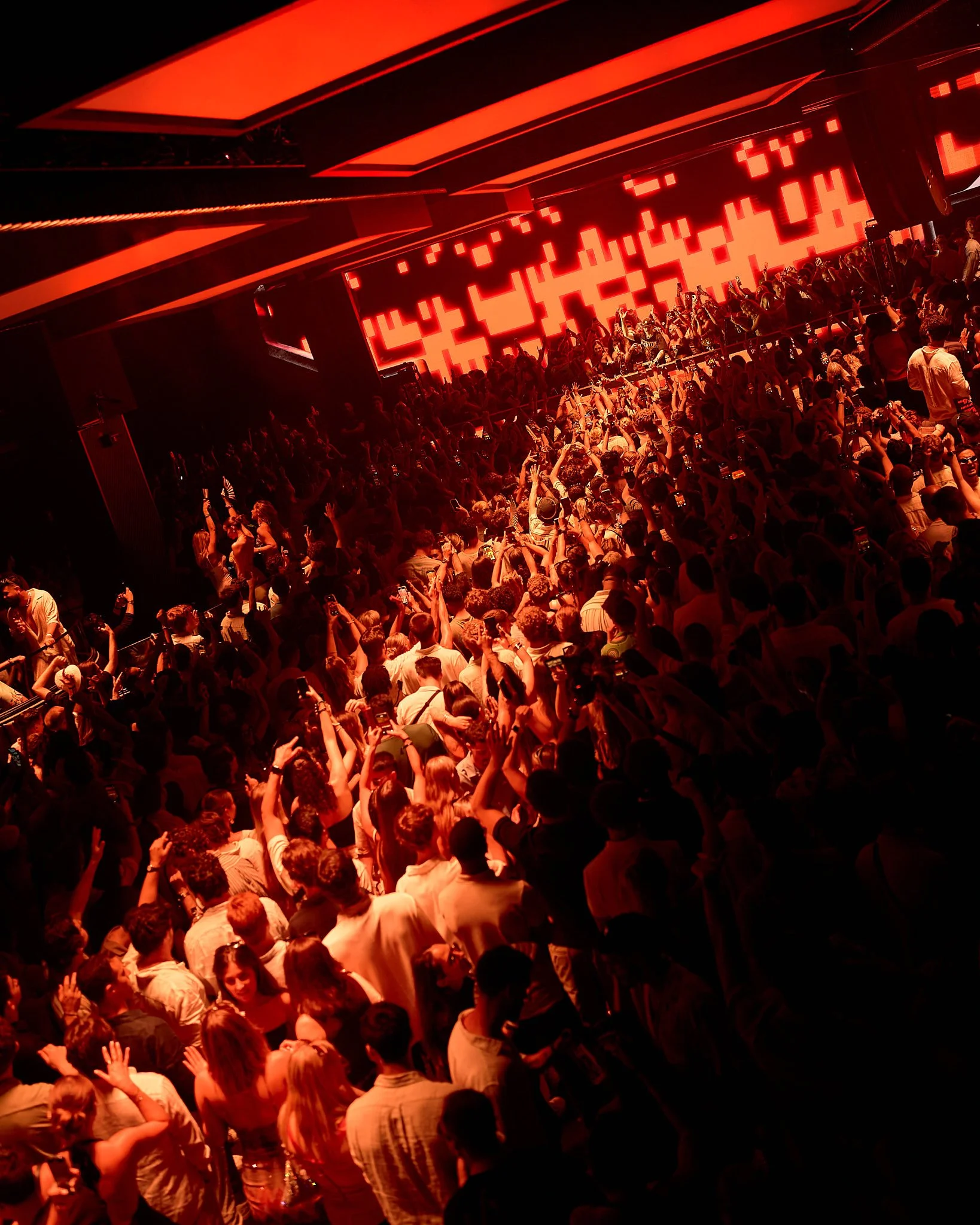 Una multitud de personas en un concierto o fiesta, con luces rojas y una pantalla grande en el fondo.