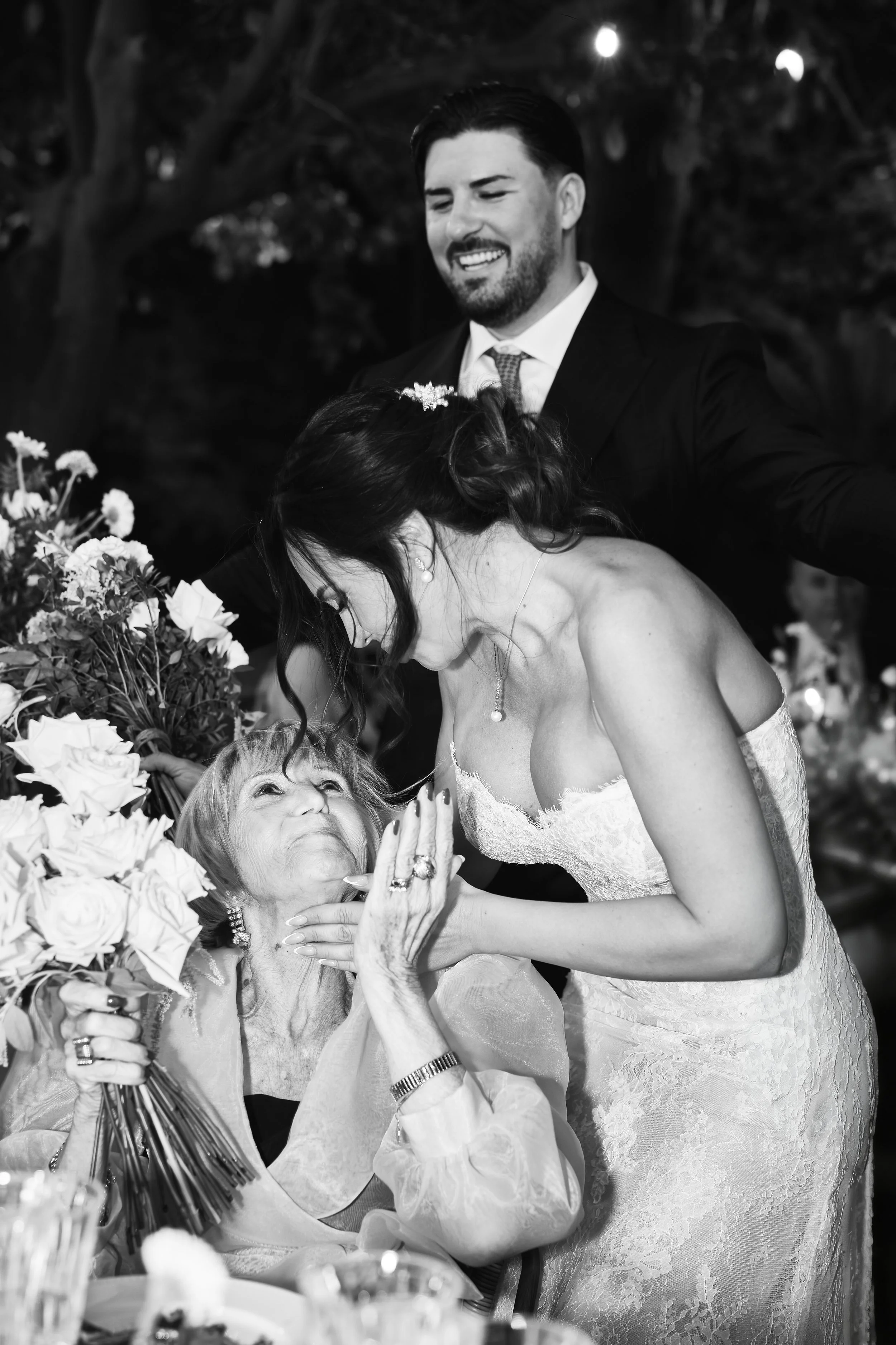 Mujer mayor con cabello rubio y vestido de novia interactuando con una mujer adulta de cabello oscuro y collar, rodeadas de flores, en un ambiente de celebración o boda.