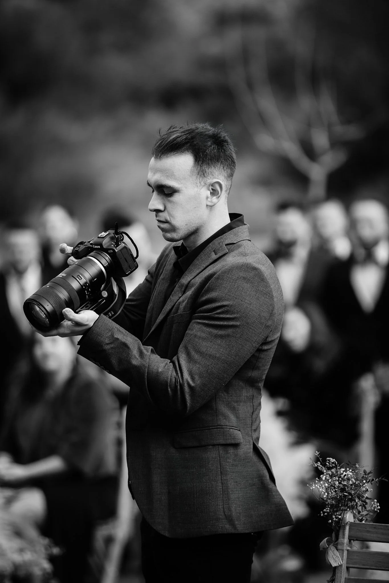 Un hombre con traje negro sostiene una cámara profesional y mira la pantalla en un entorno exterior en blanco y negro.