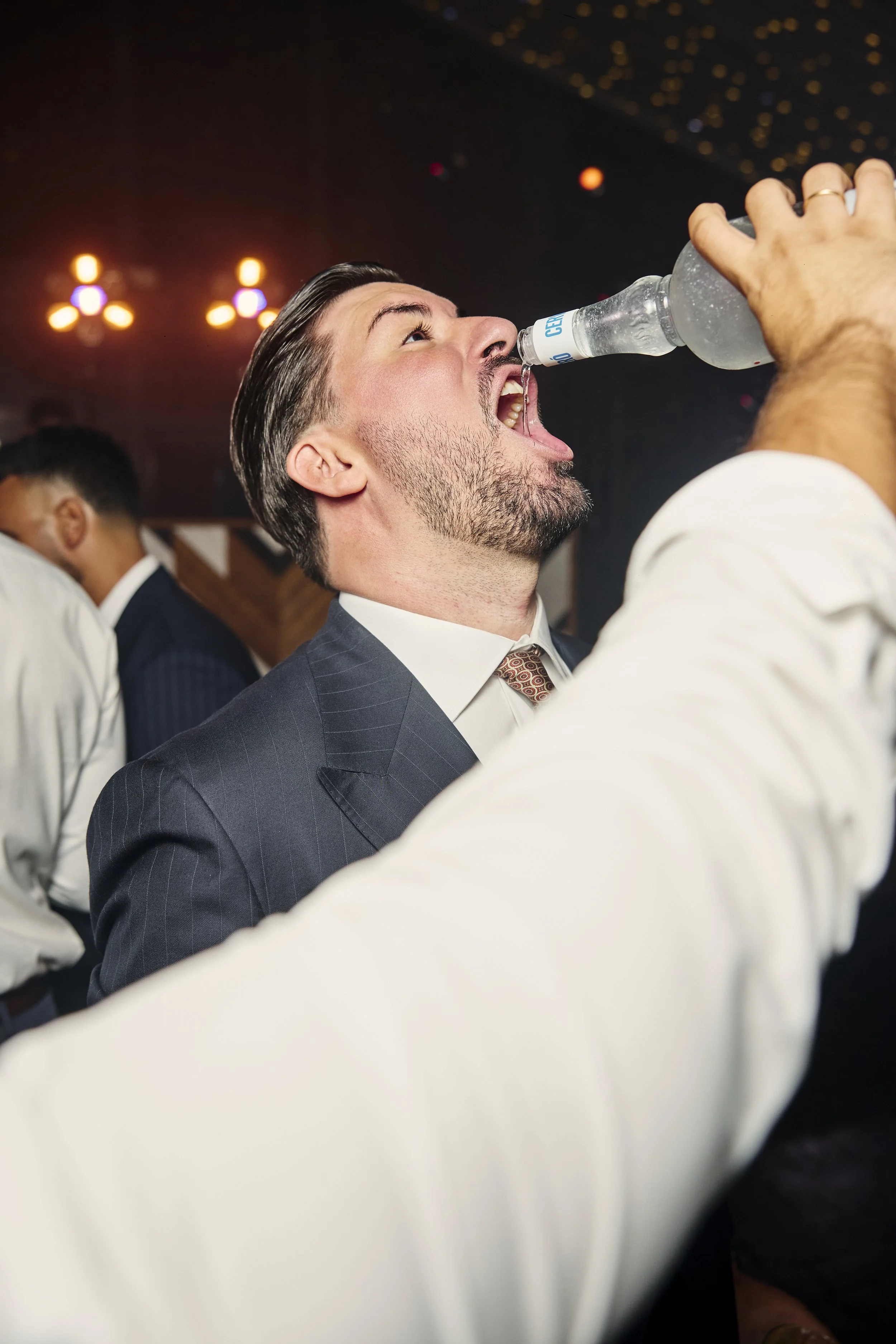 Hombre con barba y cabello peinado hacia atrás bebe agua de una botella en una celebración formal, con decoraciones de luces en el fondo.