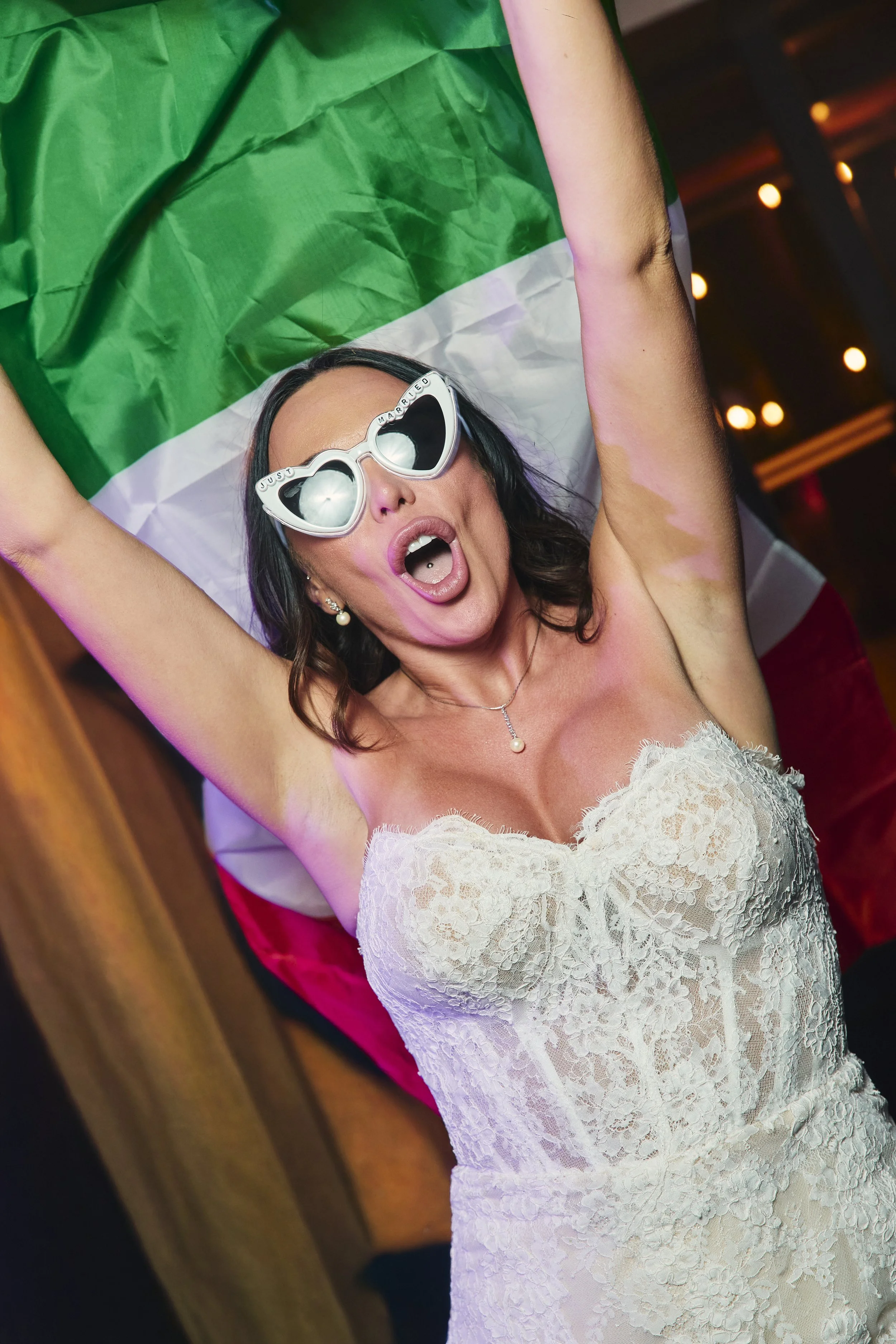 Mujer con vestido de encaje blanco, lentes de corazón con la frase "Just Mariedo", y un collar de perlas, sosteniendo una bandera que parece ser la de Italia en un ambiente de fiesta.