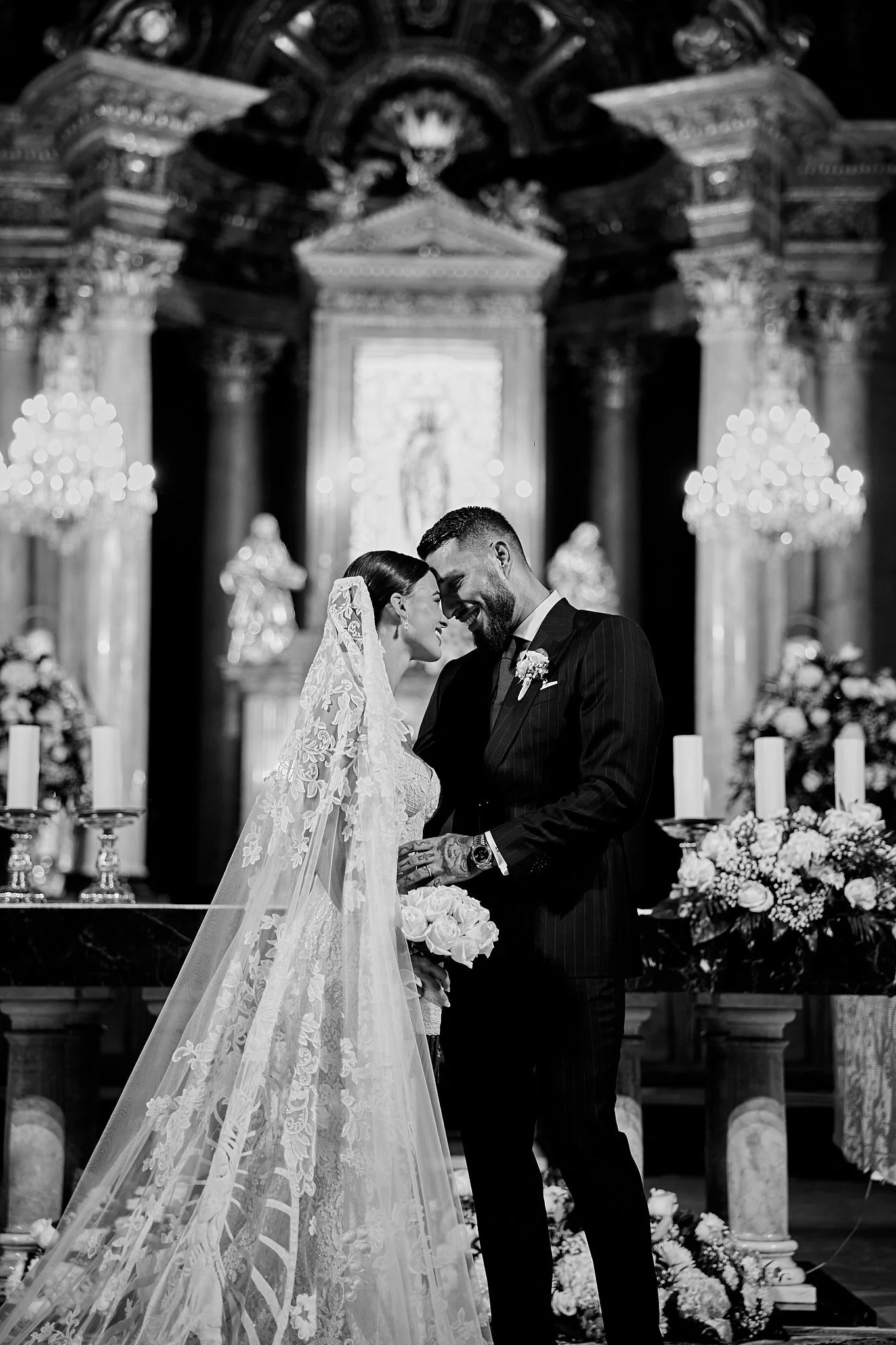 La foto muestra a una pareja de novios en su boda, sonriendo y tocándose la frente dentro de una iglesia decorada con flores y candelabros.