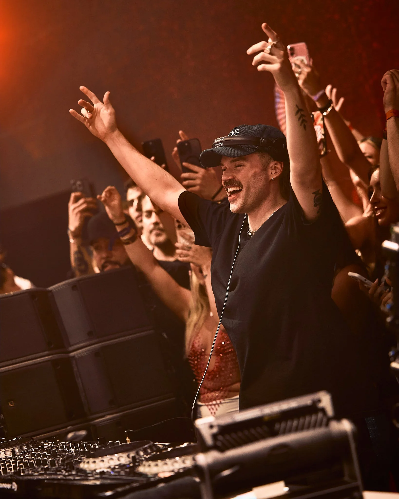 Un DJ con gorra y camiseta negra disfrutando en un concierto con una multitud de personas bailando y tomando fotos, ambiente de fiesta y música.