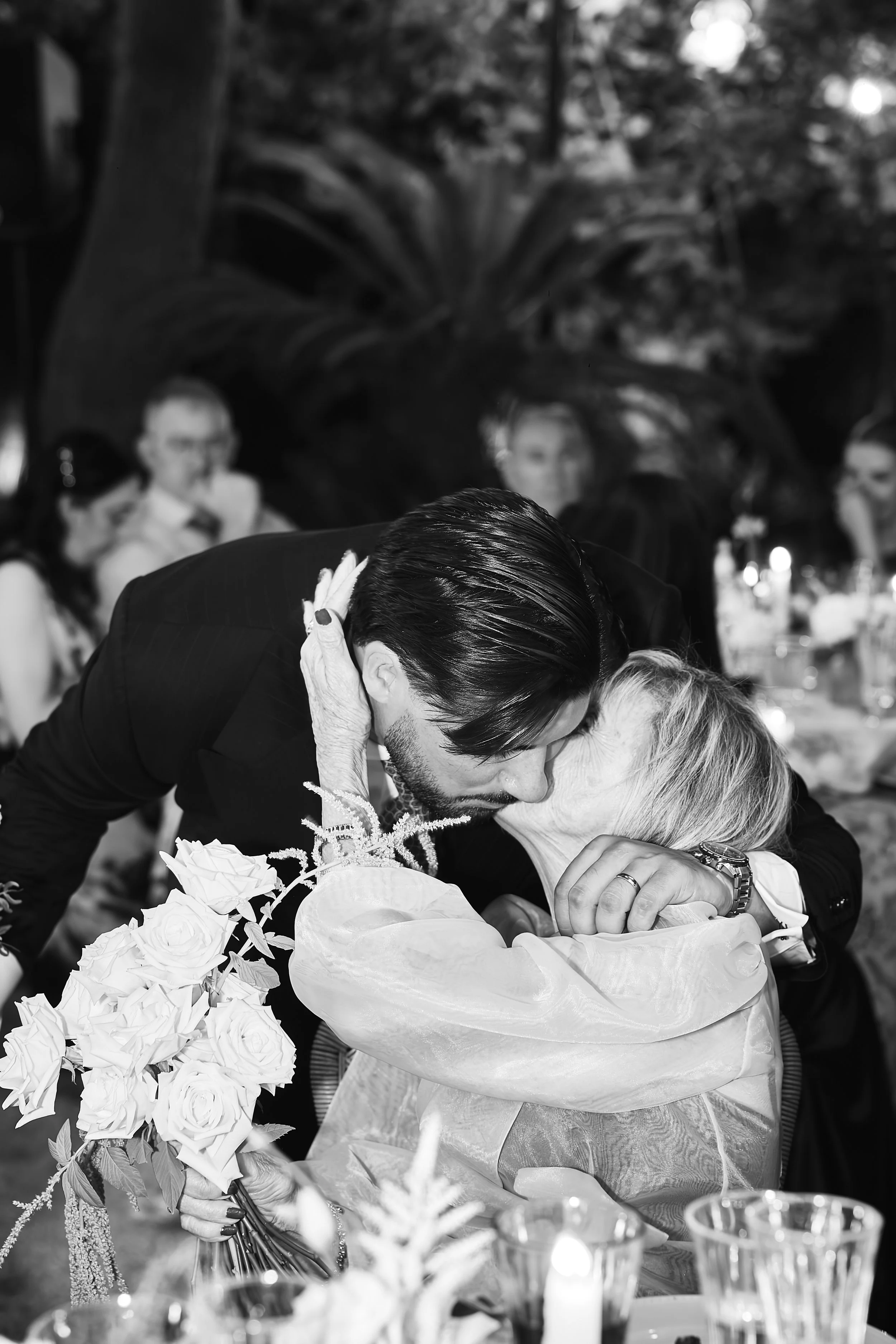 Una pareja en un evento, la mujer recibe un beso en la frente del hombre, rodeados por flores y velas.