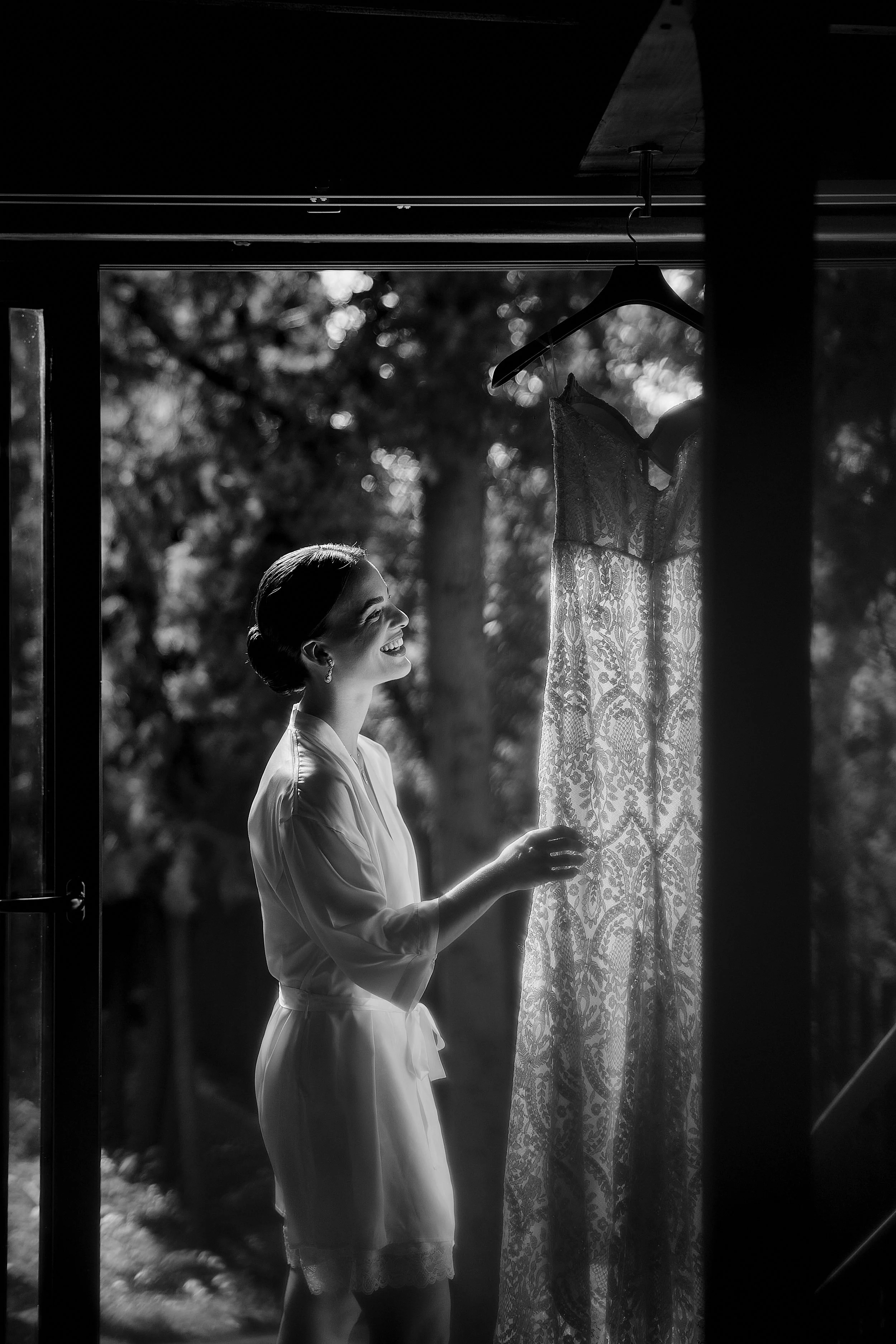 Mujer sonriendo en la ventana, mirando un vestido de encaje colgado del closet, en un ambiente al aire libre, en blanco y negro.