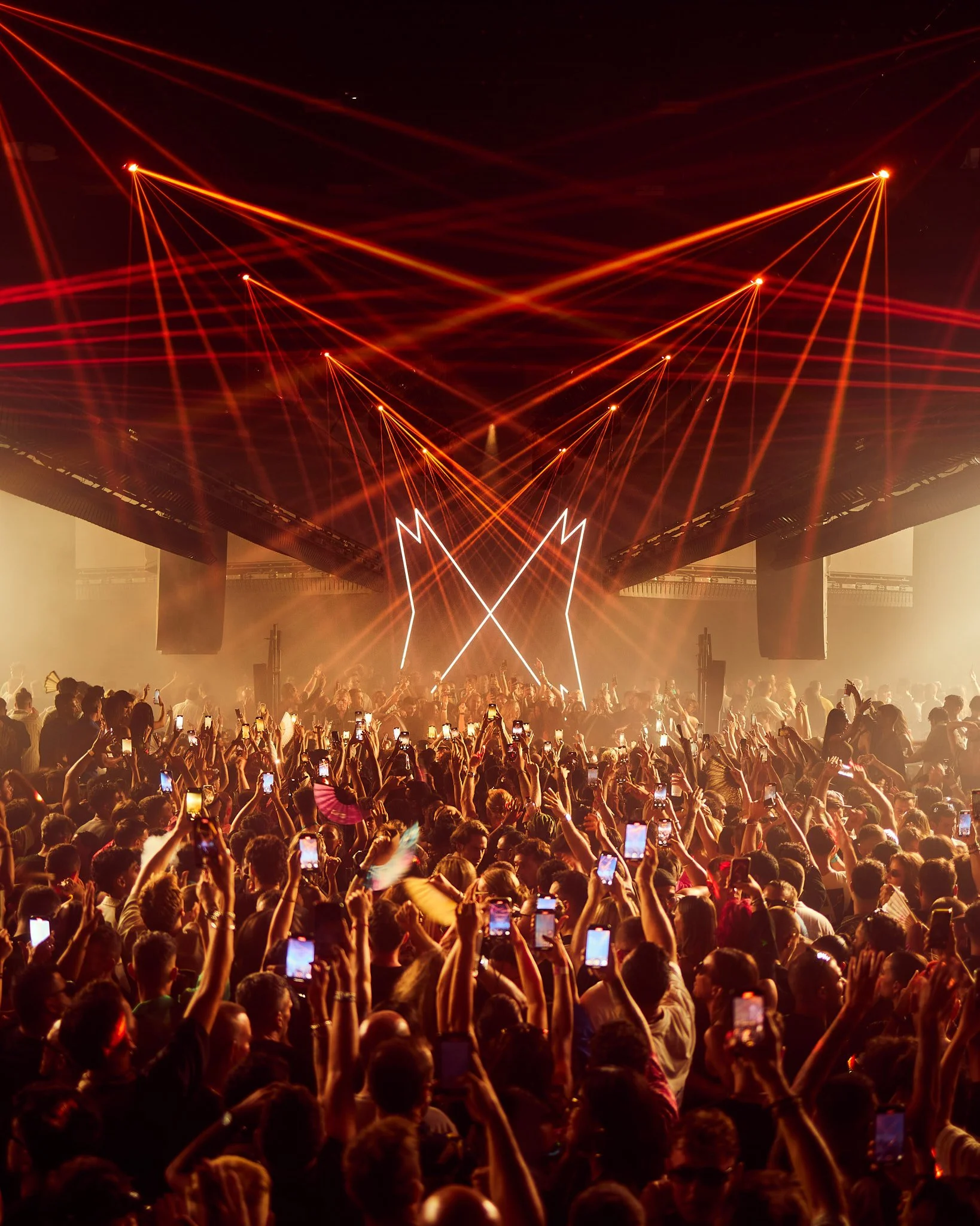 Una multitud en un concierto con luces rojas y móviles en mano, en un escenario con iluminación y efectos de láser.
