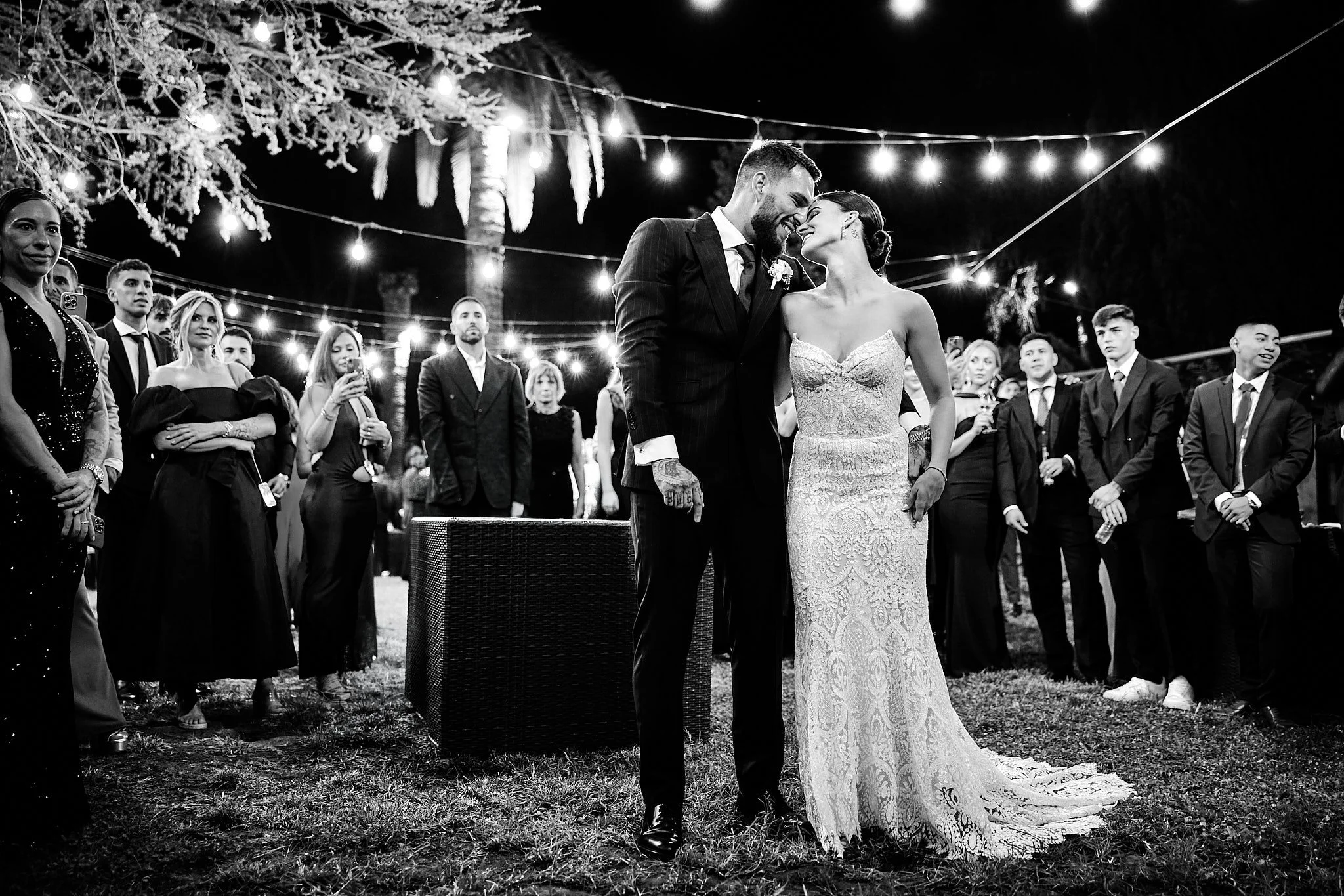 Una pareja de novios en su boda, rodeados de invitados, en una ceremonia al aire libre en la noche, con luces decorativas en el fondo.