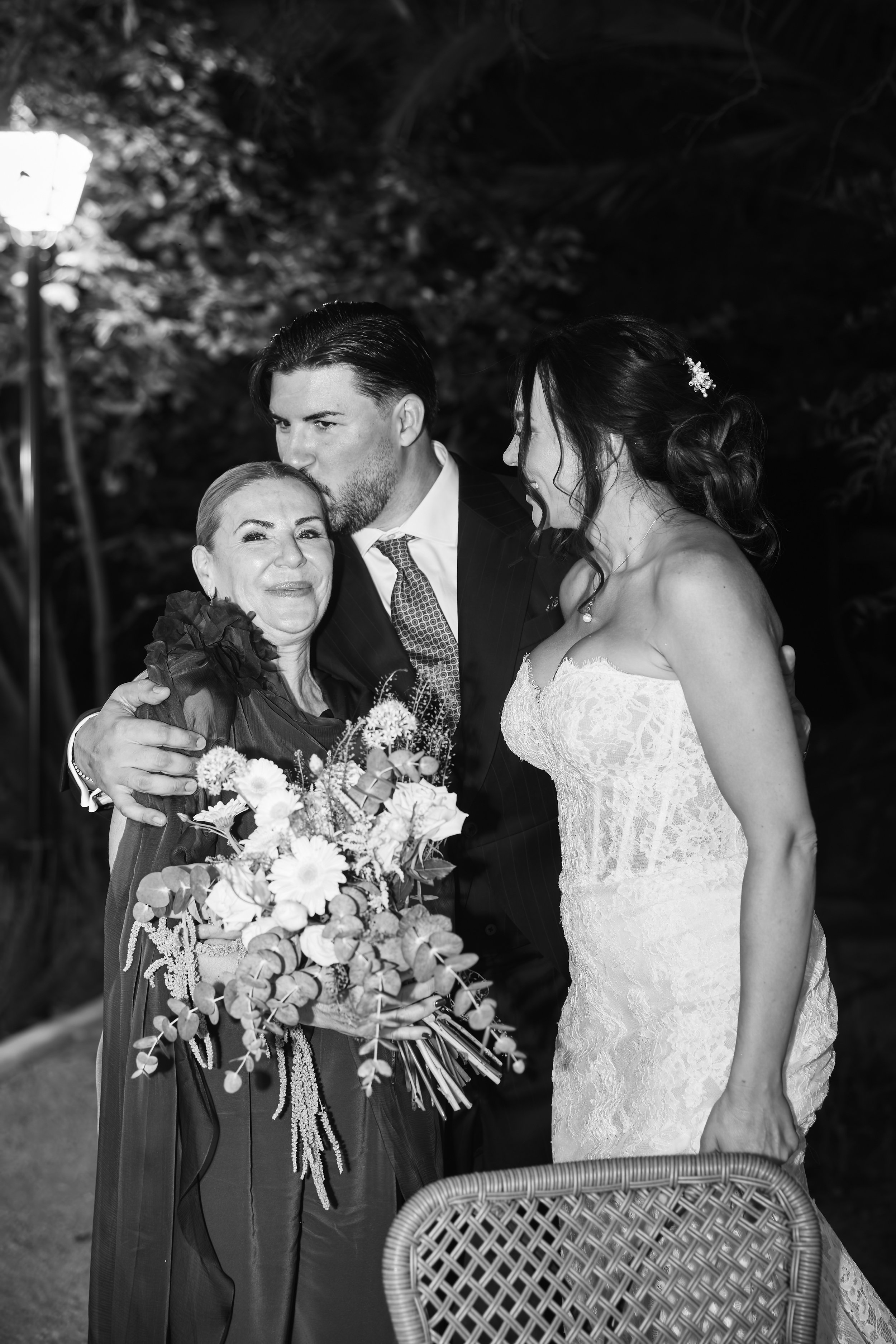 Hombre en traje abrazando a mujer en vestido de boda mientras una mujer mayor les da un beso en la frente, en un ambiente exterior de noche.