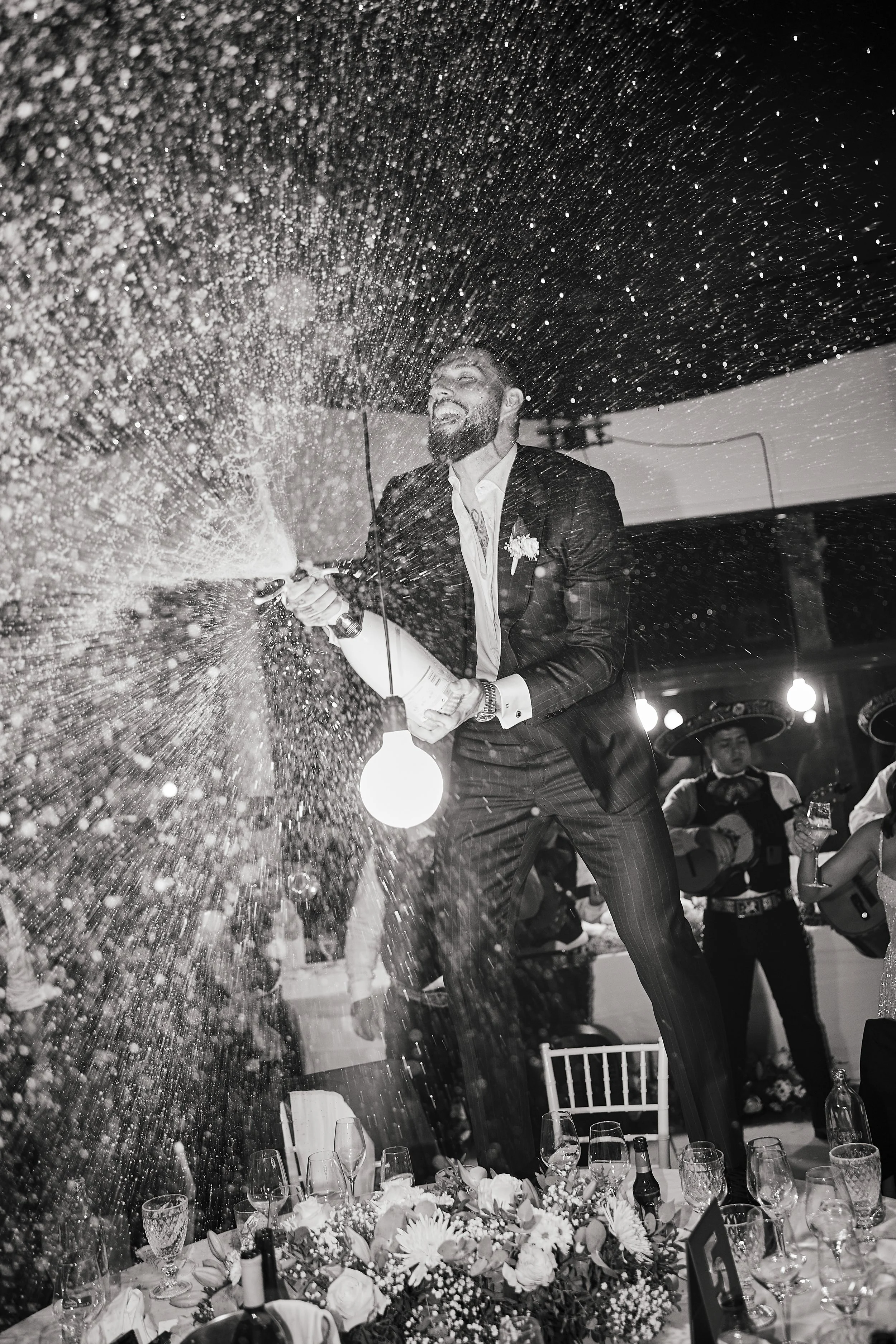 Un hombre vestido con traje celebrando en una fiesta de bodas, abriendo una botella de champagne, con brindis y música en vivo en el fondo.