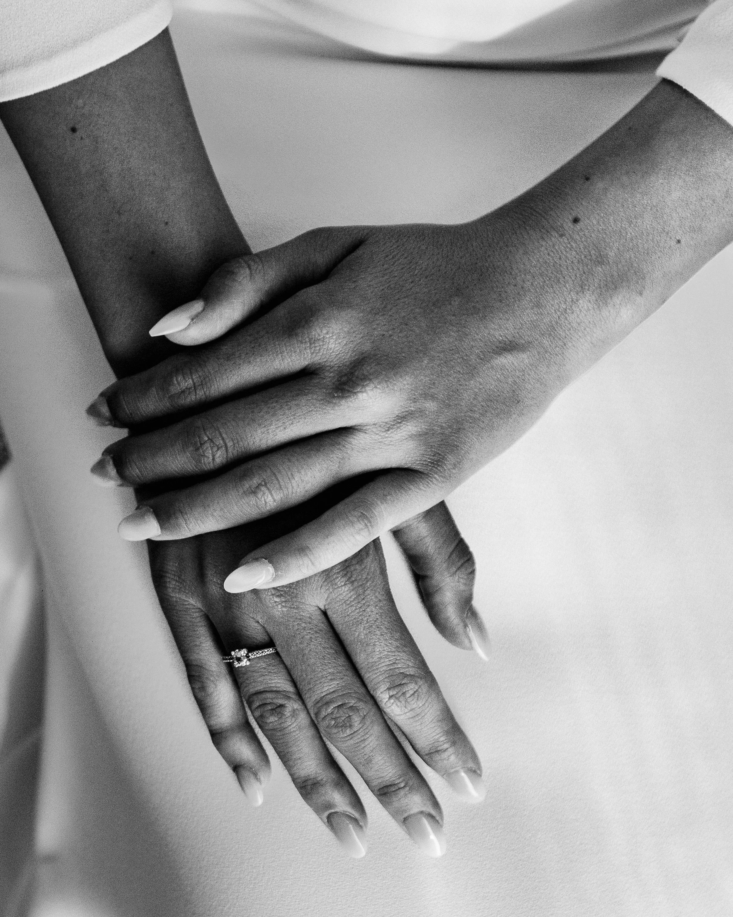 Manos de diferentes tonos de piel entrelazadas, una con un anillo de compromiso, en blanco y negro.
