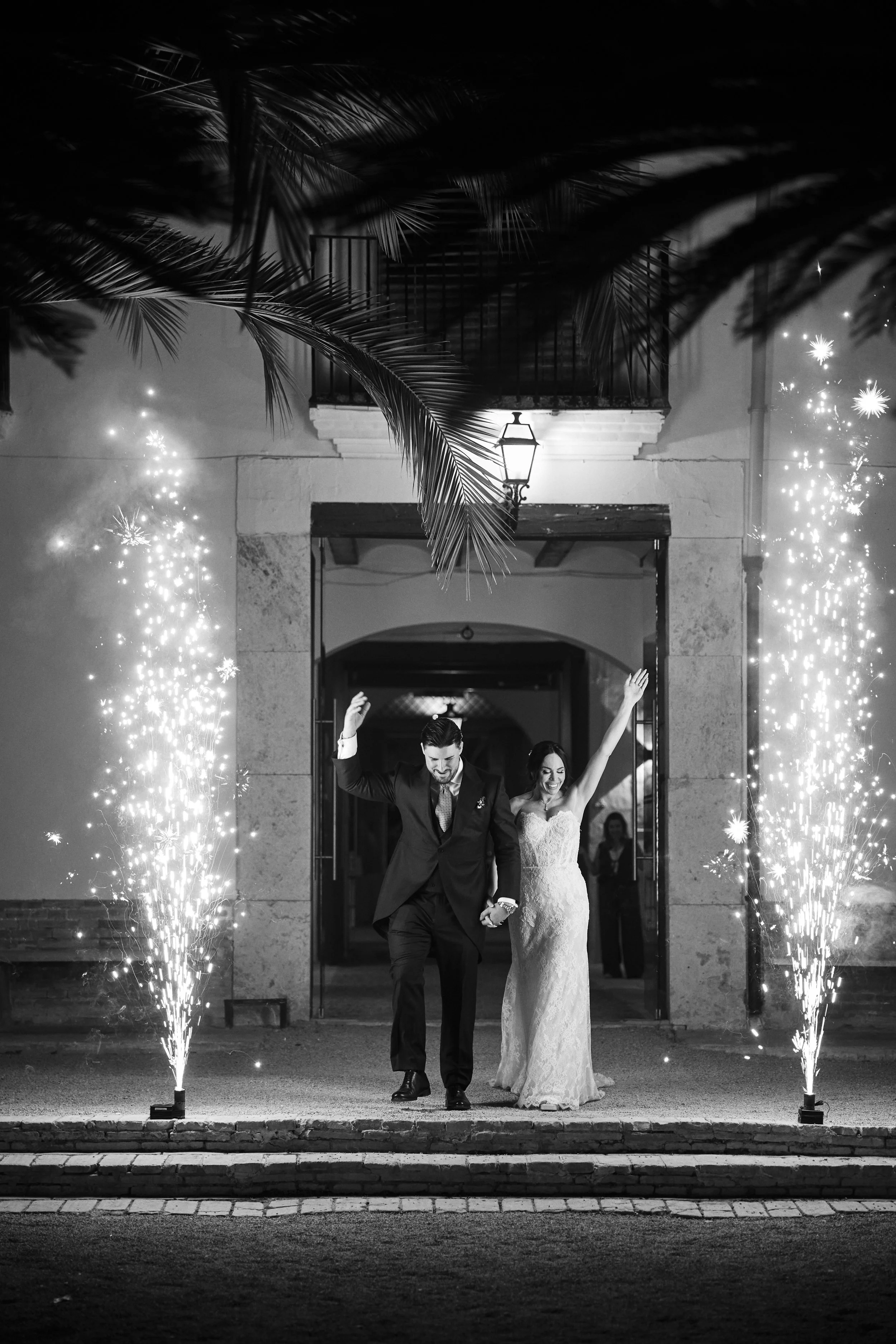 Pareja de recién casados caminando afuera de un edificio, rodeados de fuegos artificiales en la noche, con el hombre vestido en un traje negro y la mujer en un vestido de novia, sonrientes y con las manos levantadas.