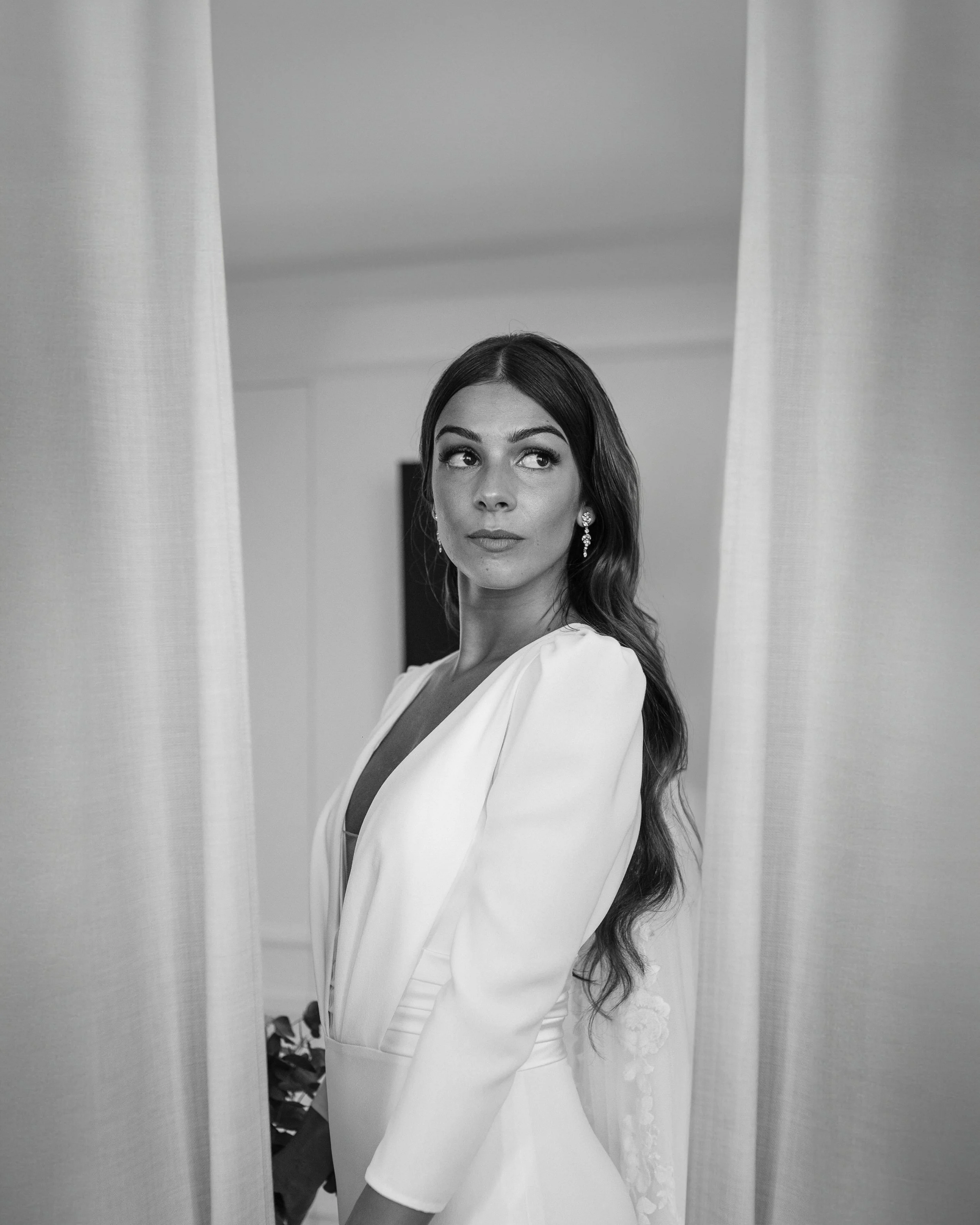 Mujer con cabello largo y rizado, vestida con un vestido blanco, observando hacia un lado, en un entorno interior.