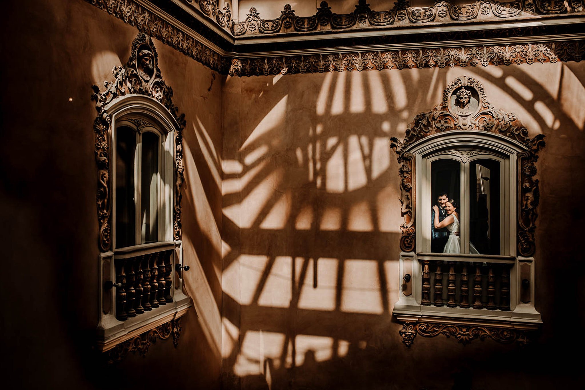 Pareja de novios sonriendo en una ventana doble en un edificio antiguo, con sombras proyectadas en la pared.