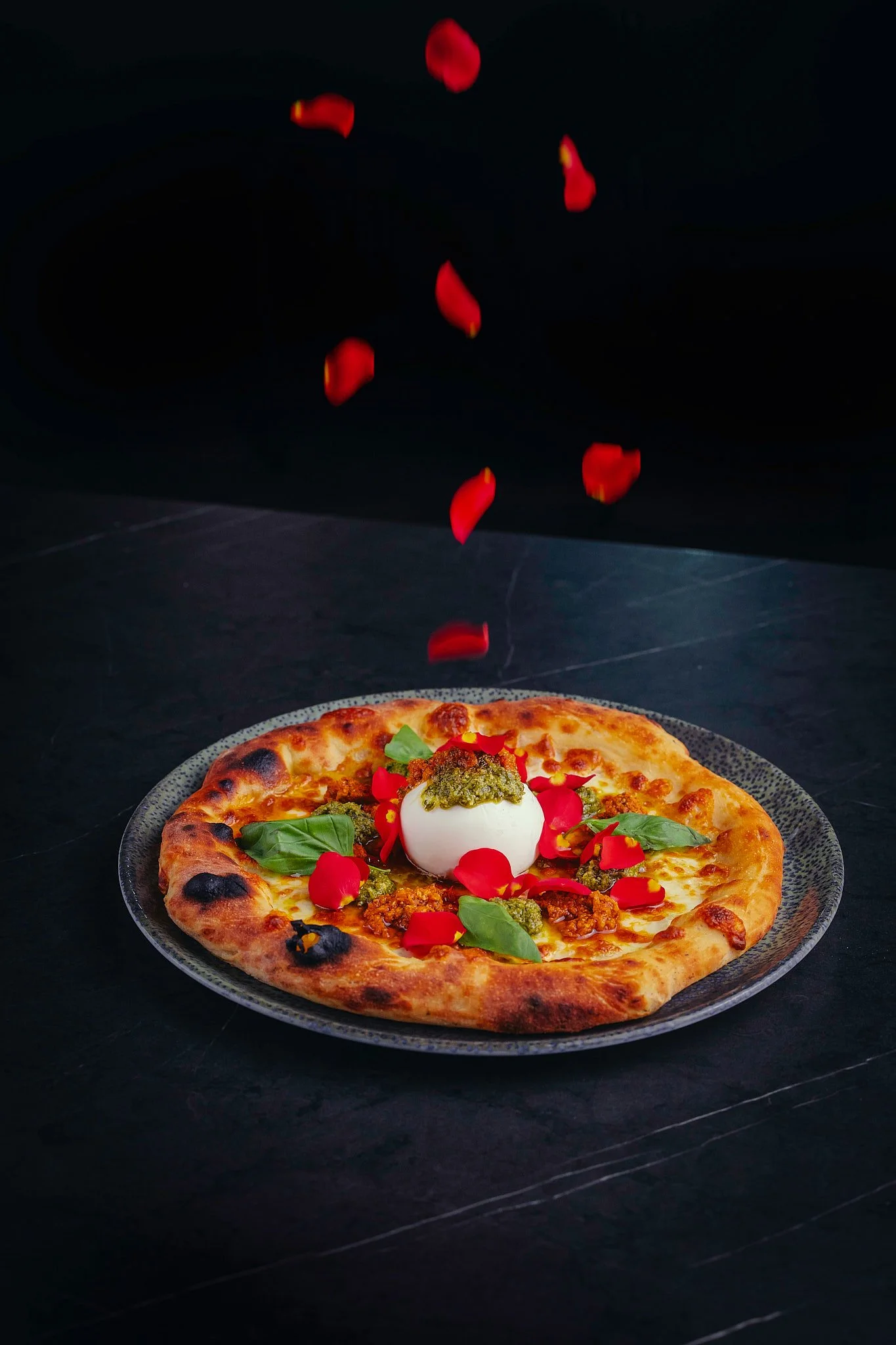 Una pizza con queso, albahaca, un huevo y hojas de albahaca decorada con pétalos de rosa.