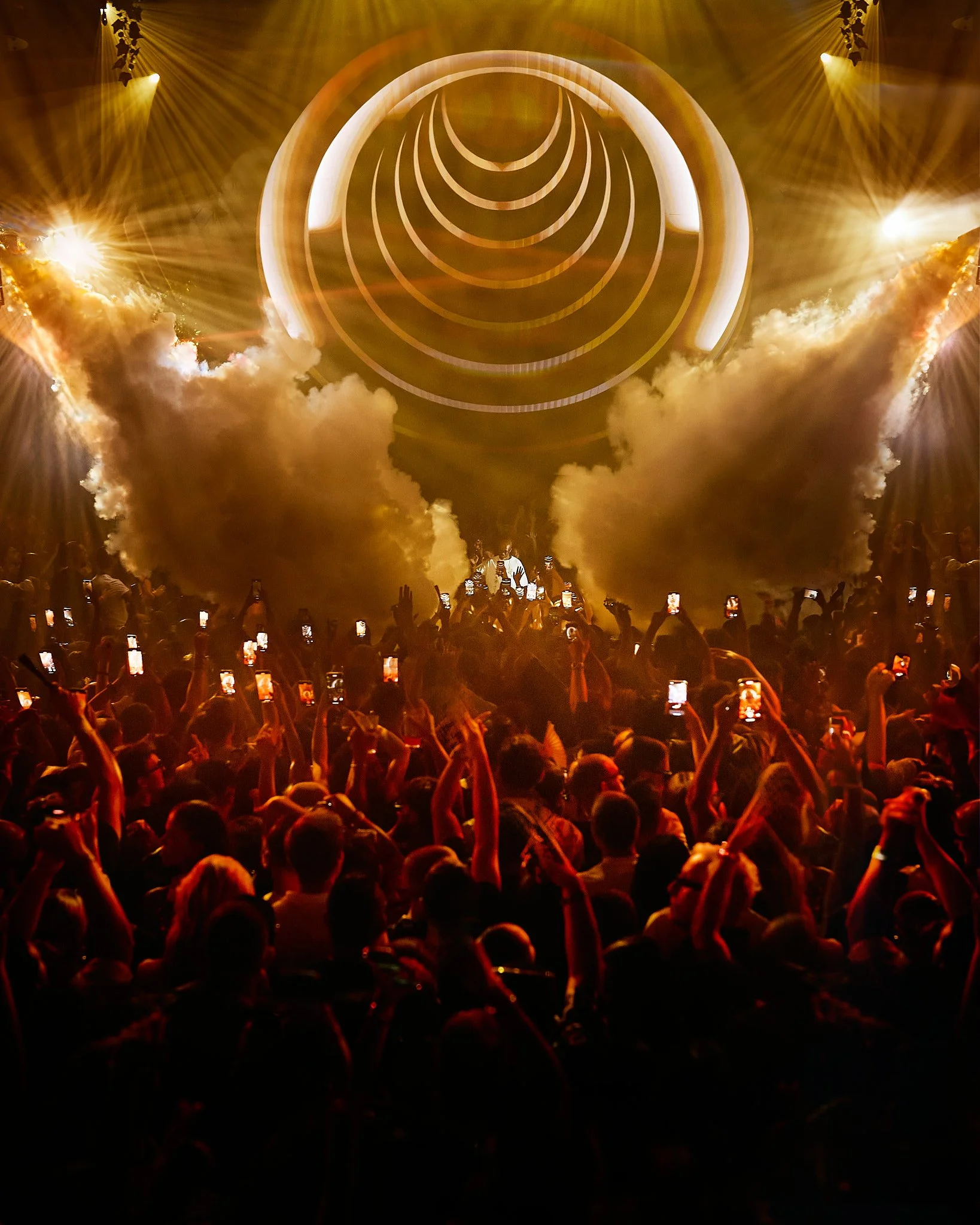Concierto con un escenario iluminado con luces doradas, un grupo grande de espectadores tomando fotos con sus teléfonos y humo en el escenario
