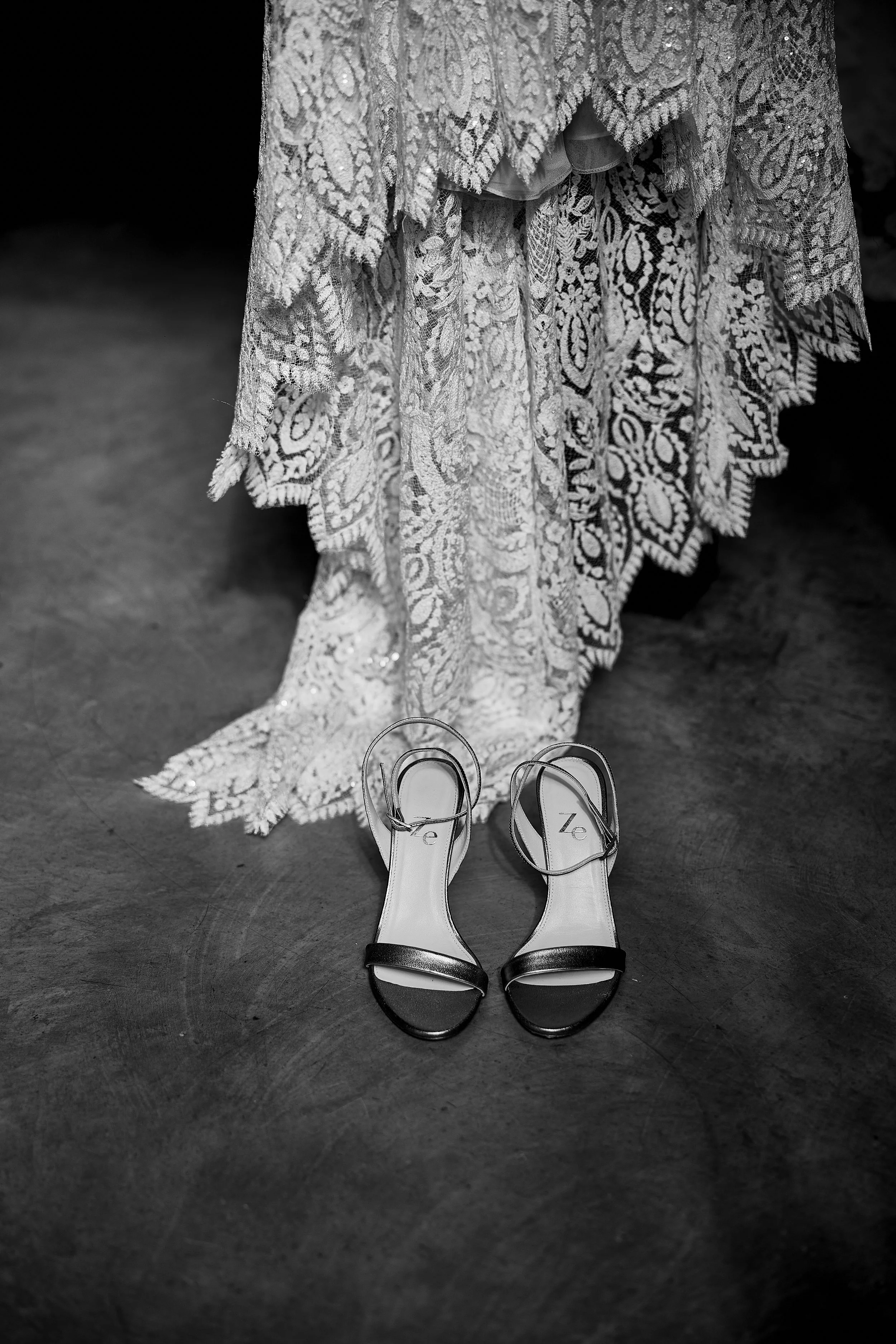 Zapatos de tacón negros y blancos en una superficie de piso, frente a un vestido de encaje. La imagen es en blanco y negro.
