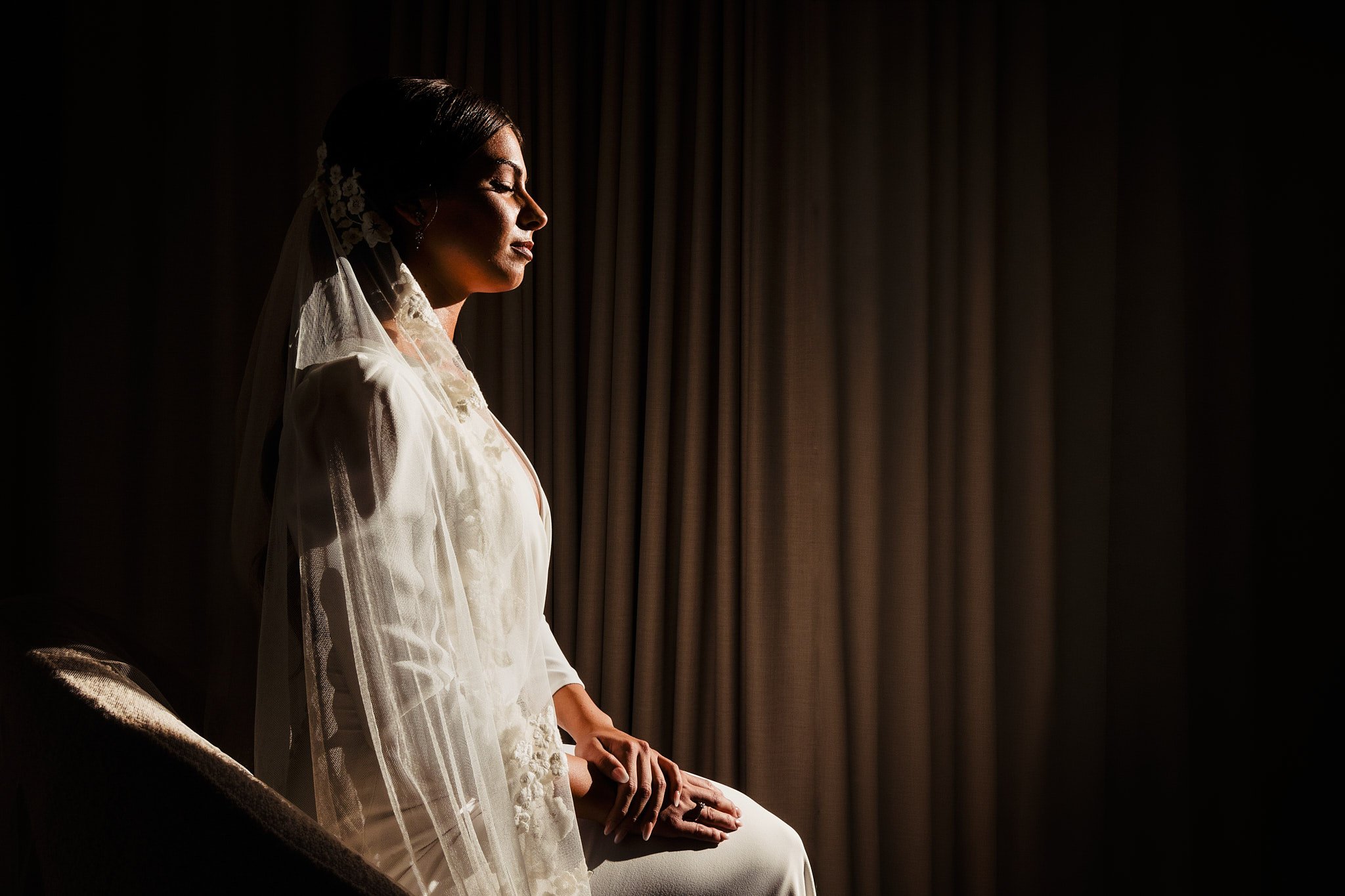 Mujer en vestido de novia con velo, sentada en una silla, en un ambiente con cortinas oscuras, iluminada por la luz de la izquierda.
