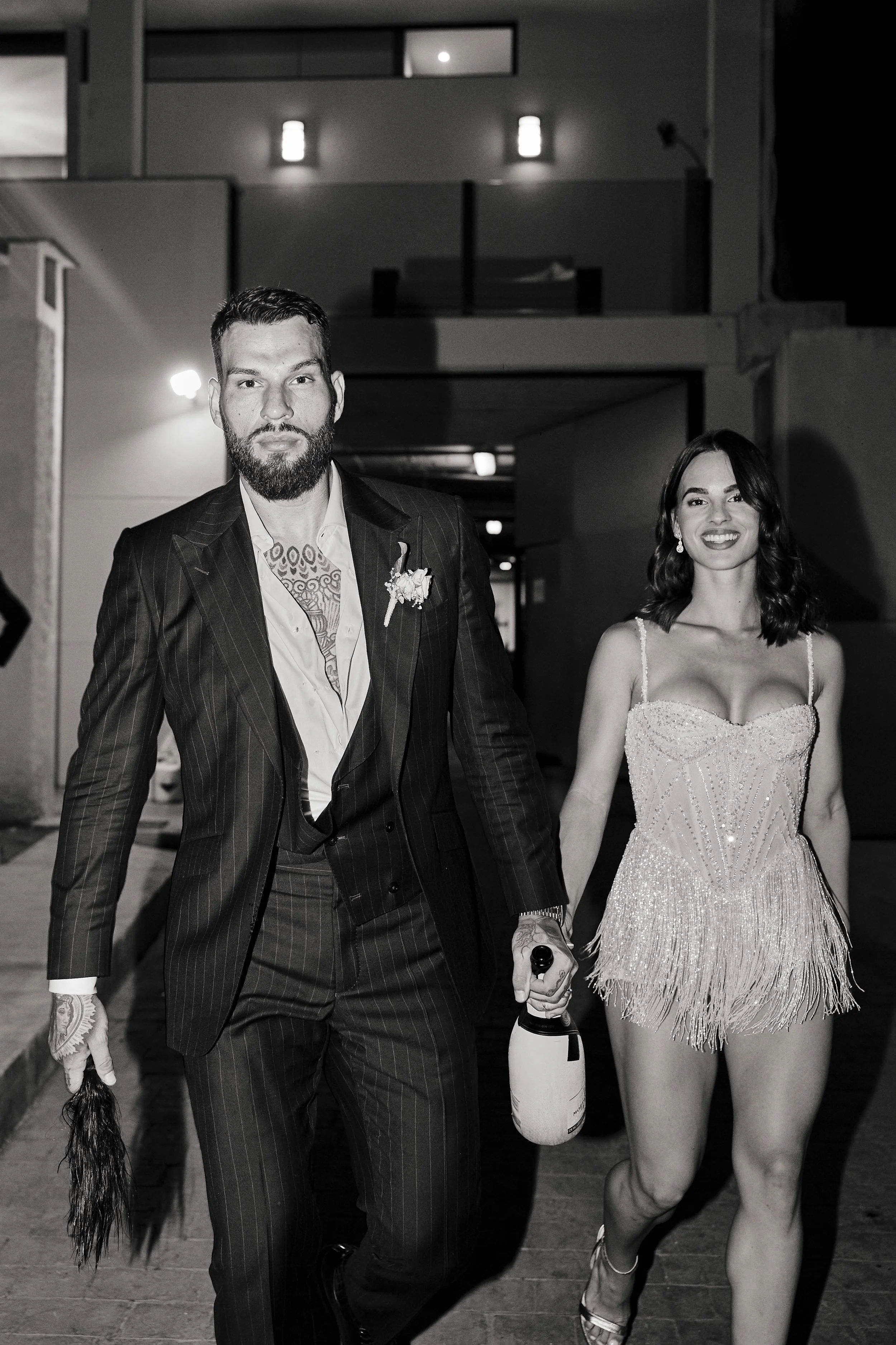 Pareja en atuendo formal caminando juntos, hombre con traje oscuro y mujer con vestido brillante, en ambiente nocturno urbano.