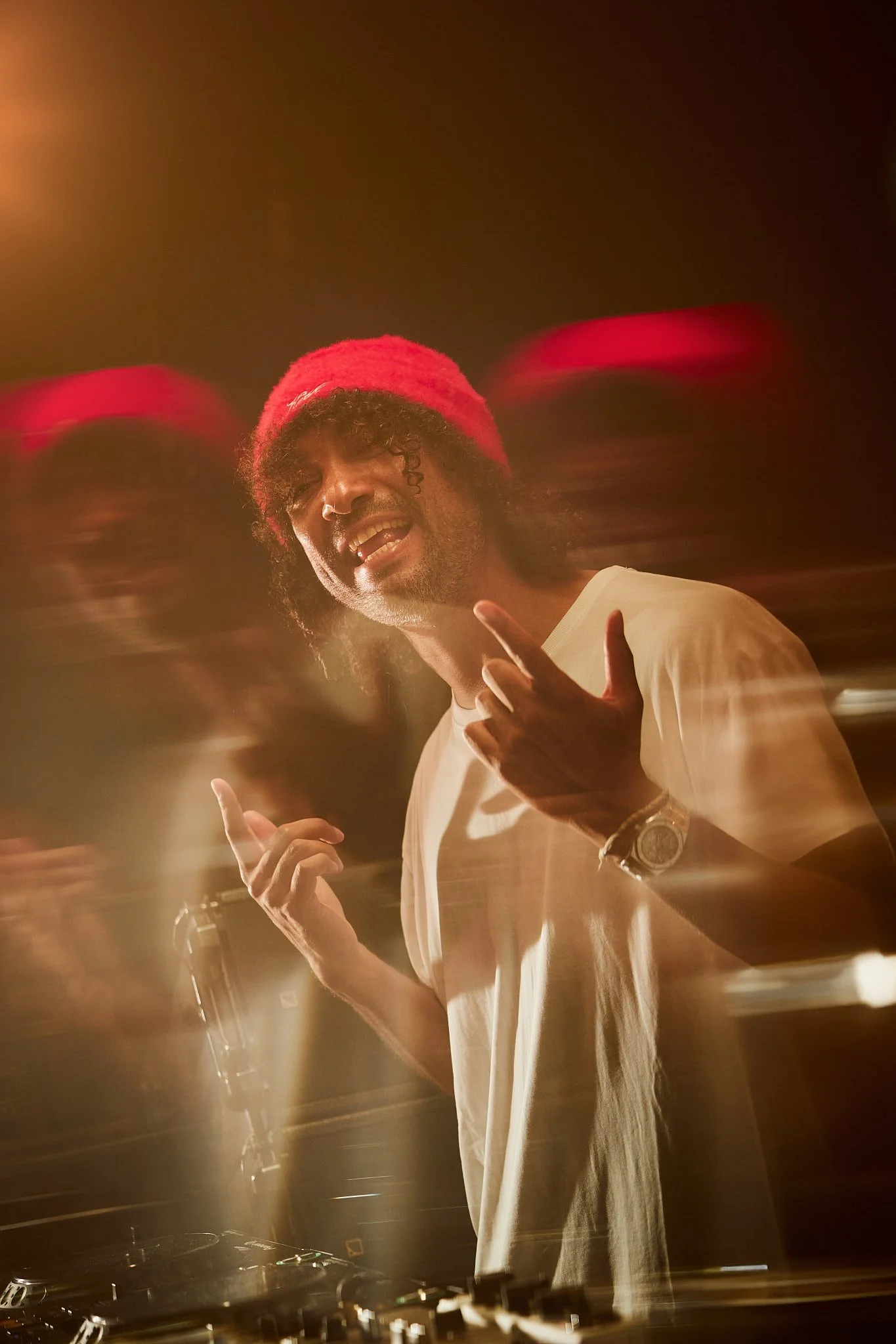 Un DJ con gorro rojo y camiseta blanca, tocando en la pista, con efectos de luz y movimiento en el fondo.