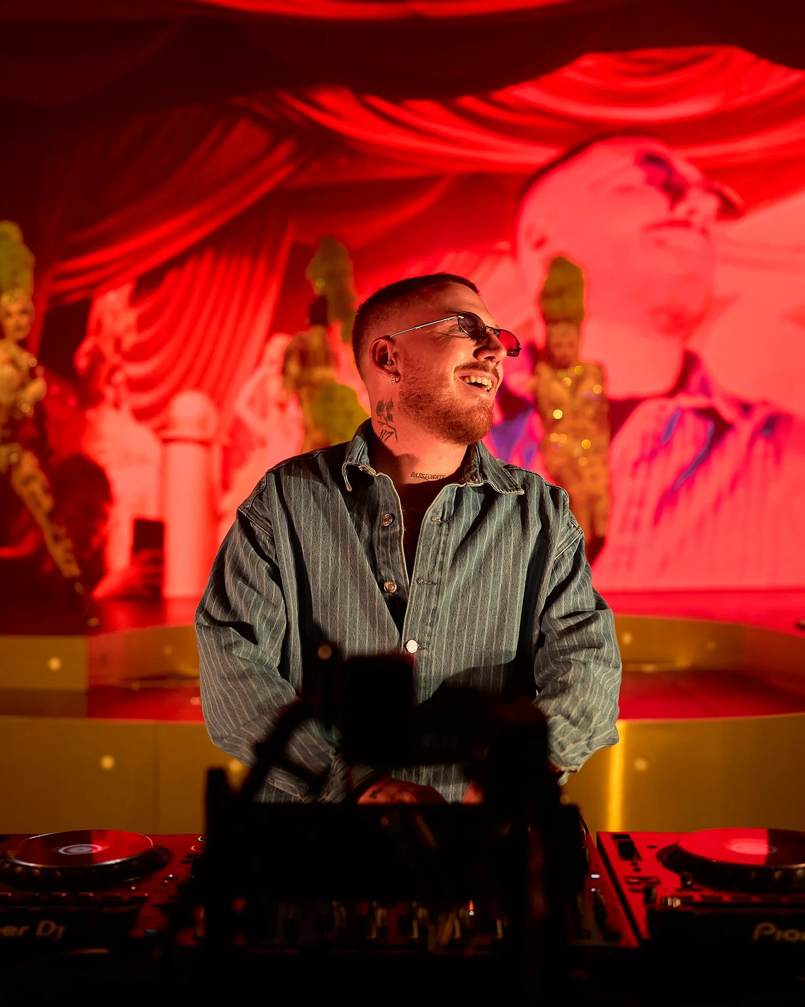 Un joven DJ con gafas de sol y camiseta de mezclilla, sonriendo frente a una mesa de DJ en un escenario con fondo rojo y figuras de artistas.