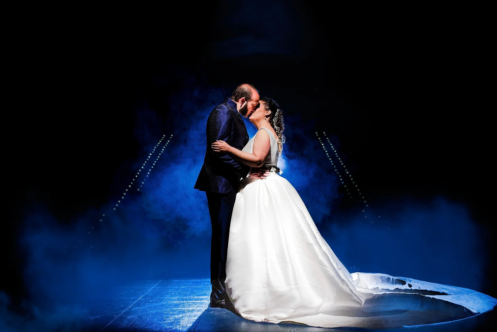 Pareja de boda abrazada y mirándose en escenario con efectos de humo azul y luces oscuras.