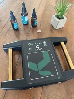 A digital foot massager or ultrasound therapy device on a wooden surface with three small bottles of oil or massage serum, a teardrop-shaped object, and a small potted plant nearby.
