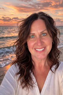 A woman with wavy brown hair and blue eyes smiling at the camera at the beach during sunset.