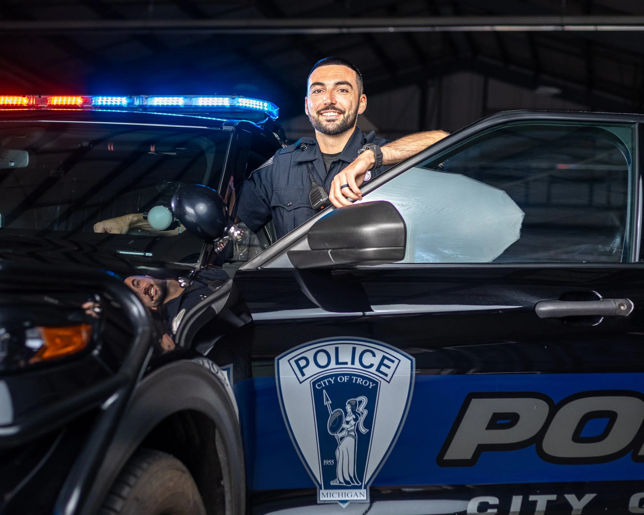 A police officer leaning on the open door of a city police car inside a garage, with flashing red and blue lights on top of the vehicle.