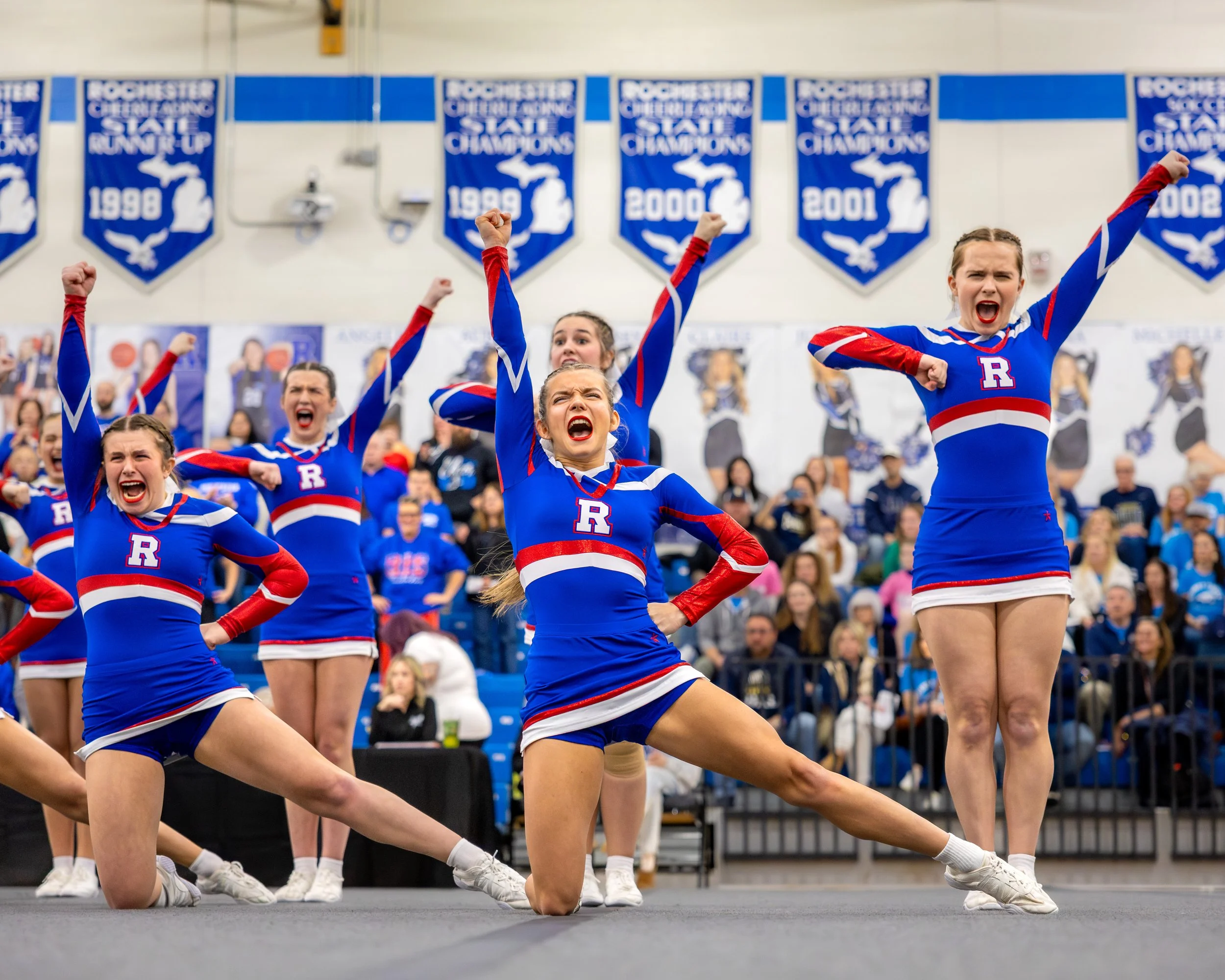 Cheerleaders performing a cheer routine at a sports event, wearing blue, red, and white uniforms with the letter 'R', in front of an audience and banners celebrating championship wins.
