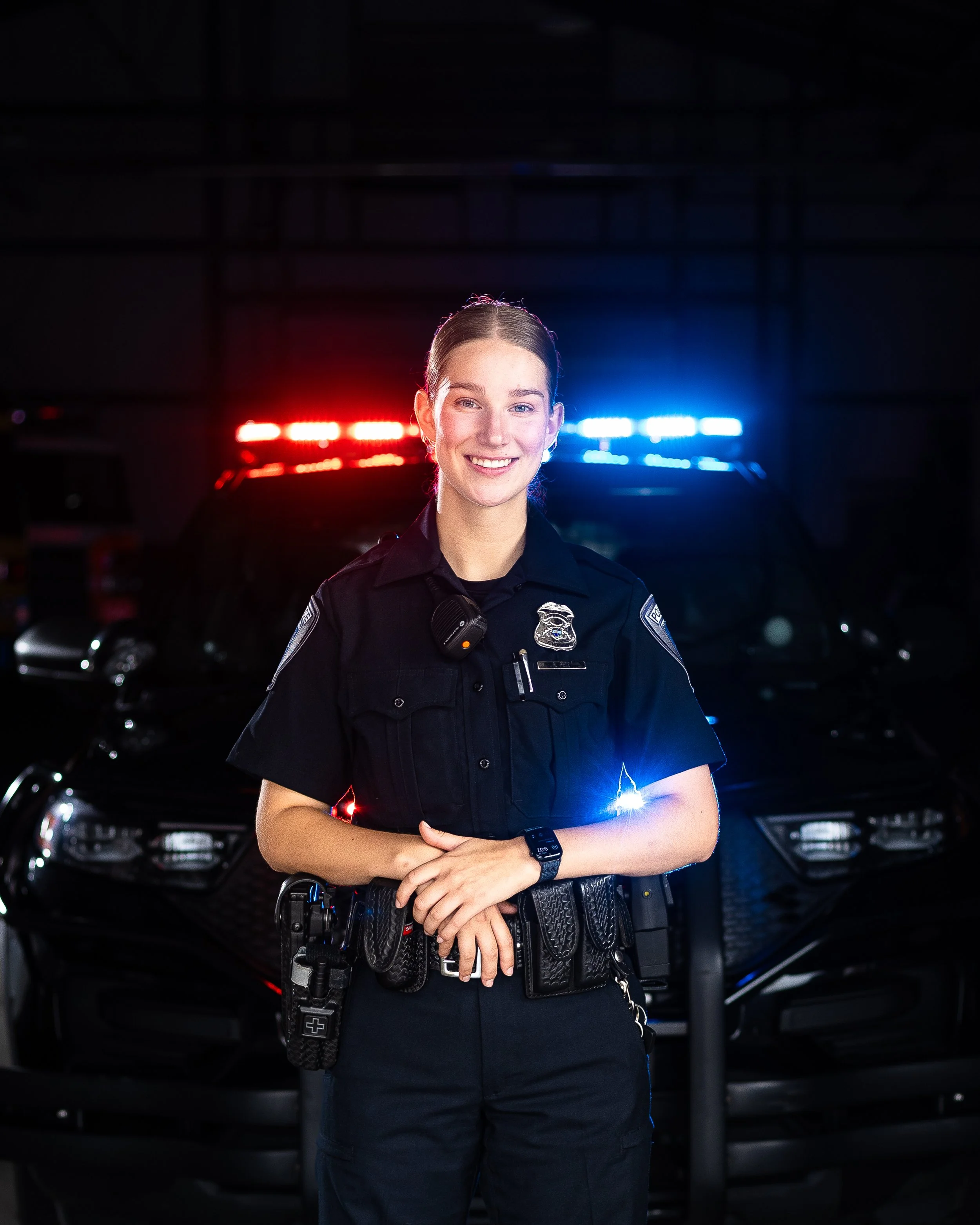 A female police officer standing in front of police cars with flashing lights.