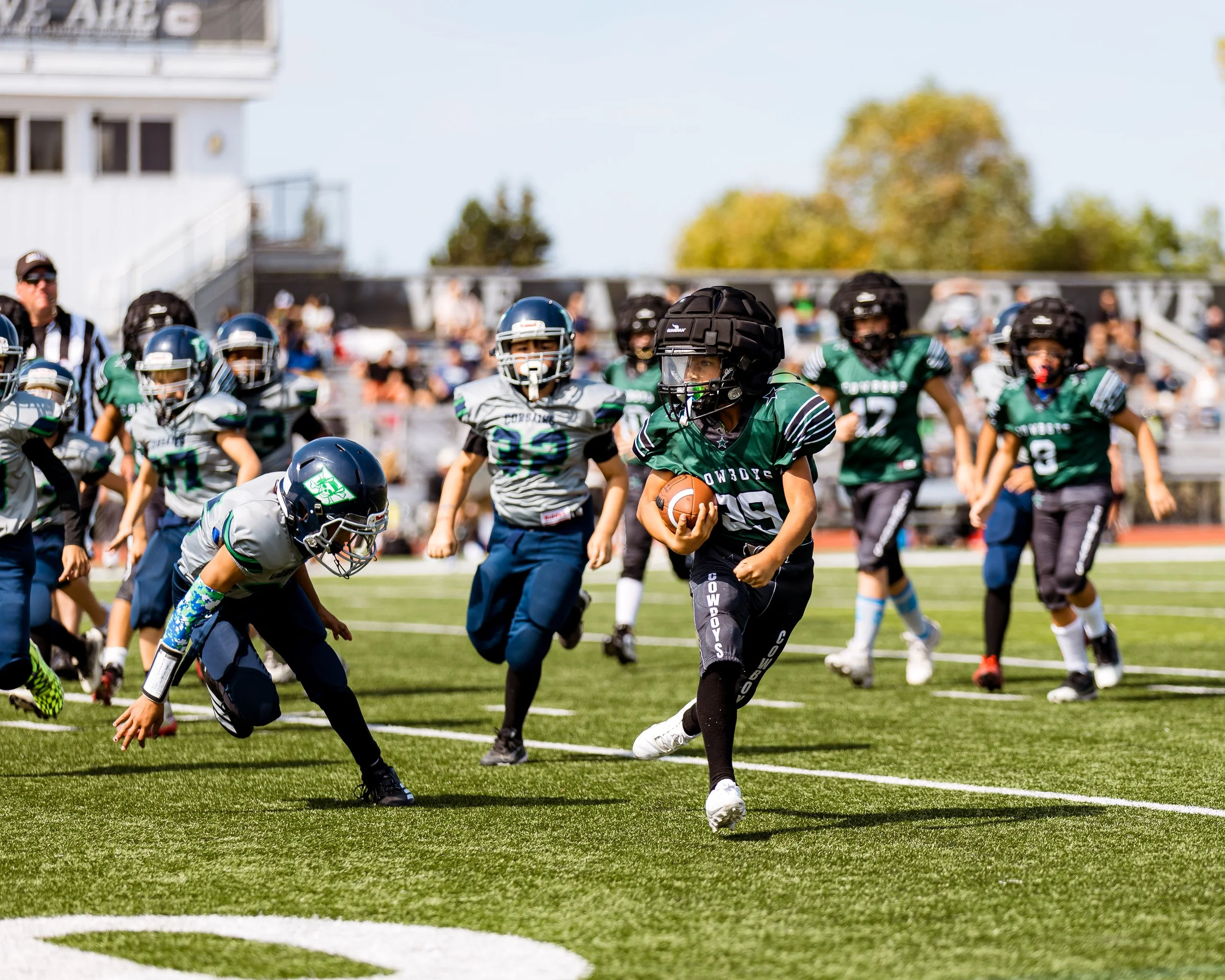 Young football players playing on a sunny day during a game, with one player in a green uniform running with the ball while others in gray uniforms pursue.