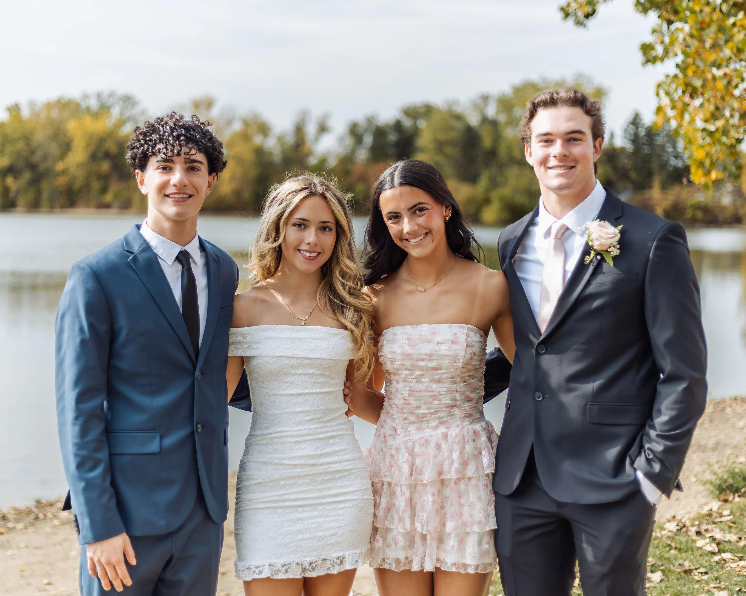 Group of four young adults dressed in formal attire, standing outdoors near a lake with trees in the background, smiling at the camera.