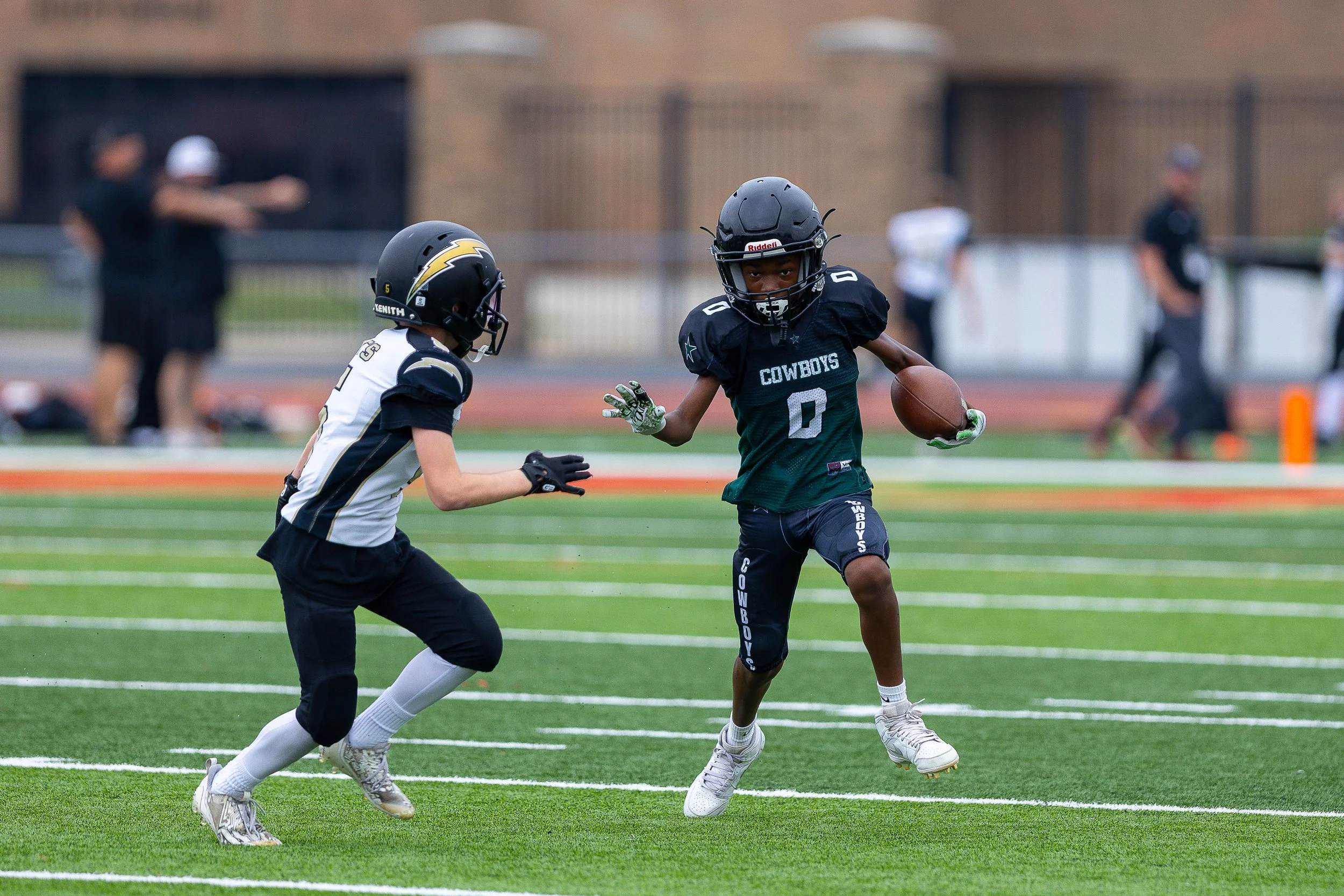 Two young football players, one in a black and white uniform and helmet, the other in a black uniform with a helmet, are on a football field. The player in black is running with the football, trying to evade the defender.