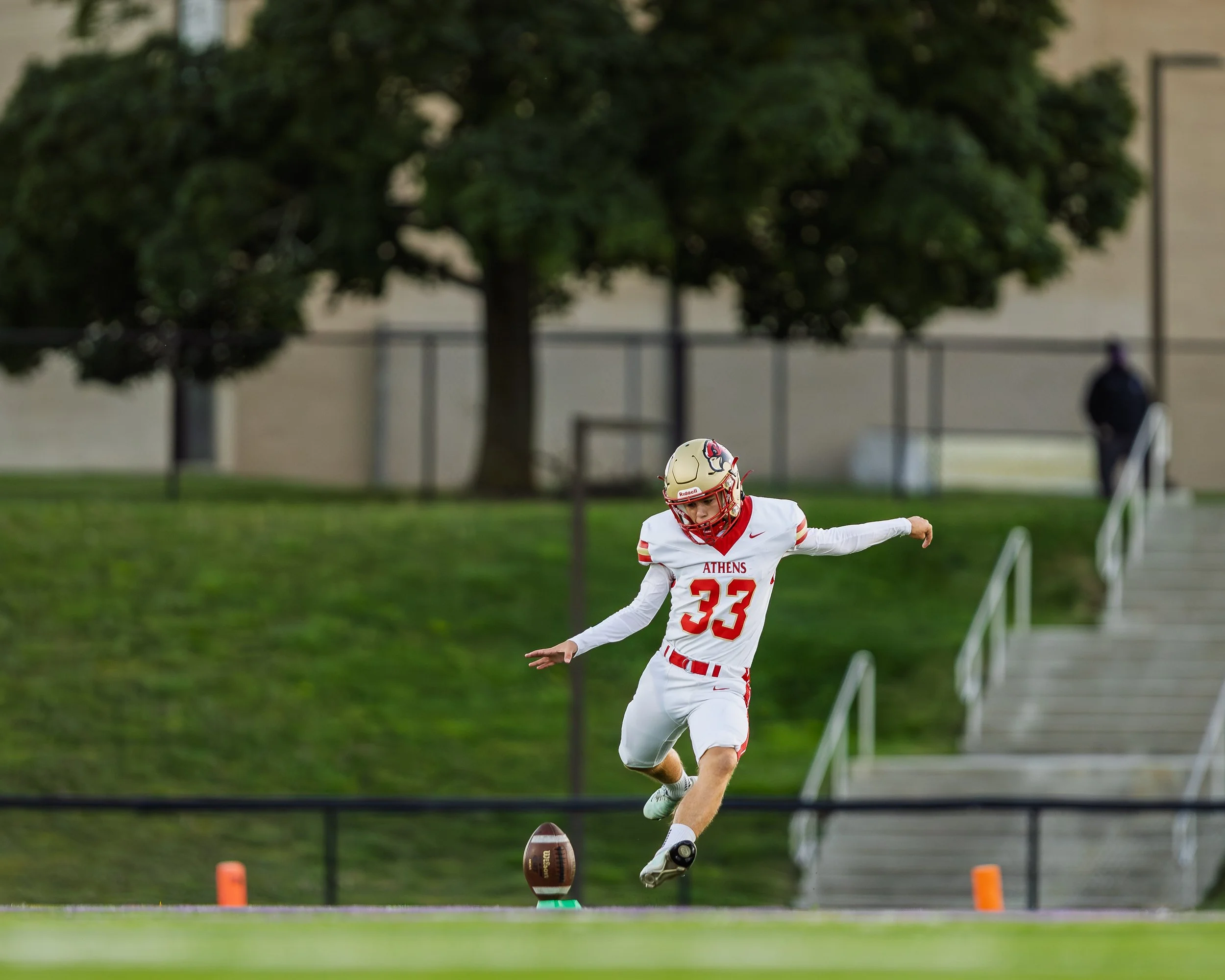 A young football player in a white and red uniform with the number 33, from Athens, is kicking a football on a field during a game or practice. There are trees and a stadium wall in the background.
