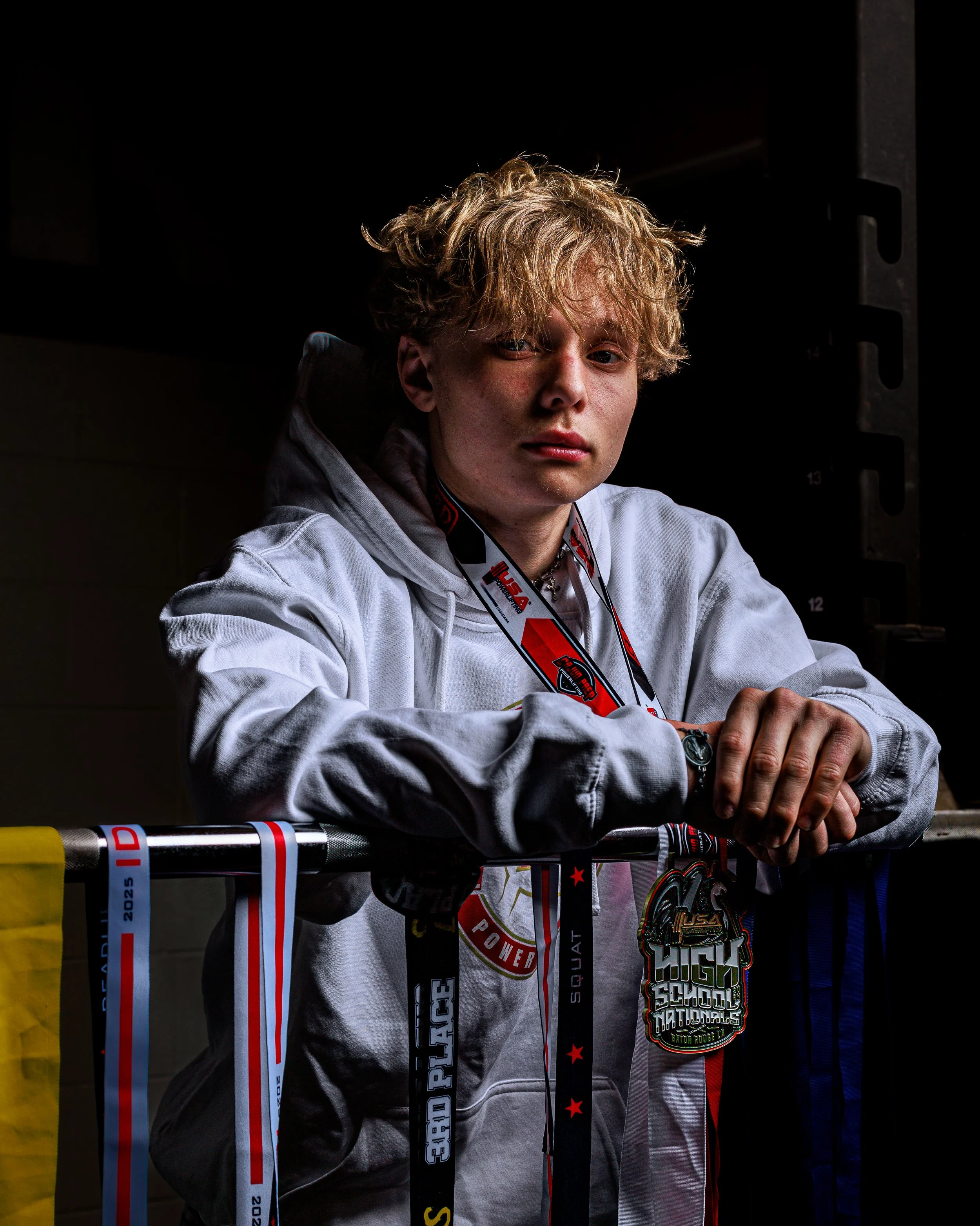 Young man with blonde curly hair, wearing a white hoodie, leaning on a railing adorned with medals and ribbons, in a dark indoor setting.
