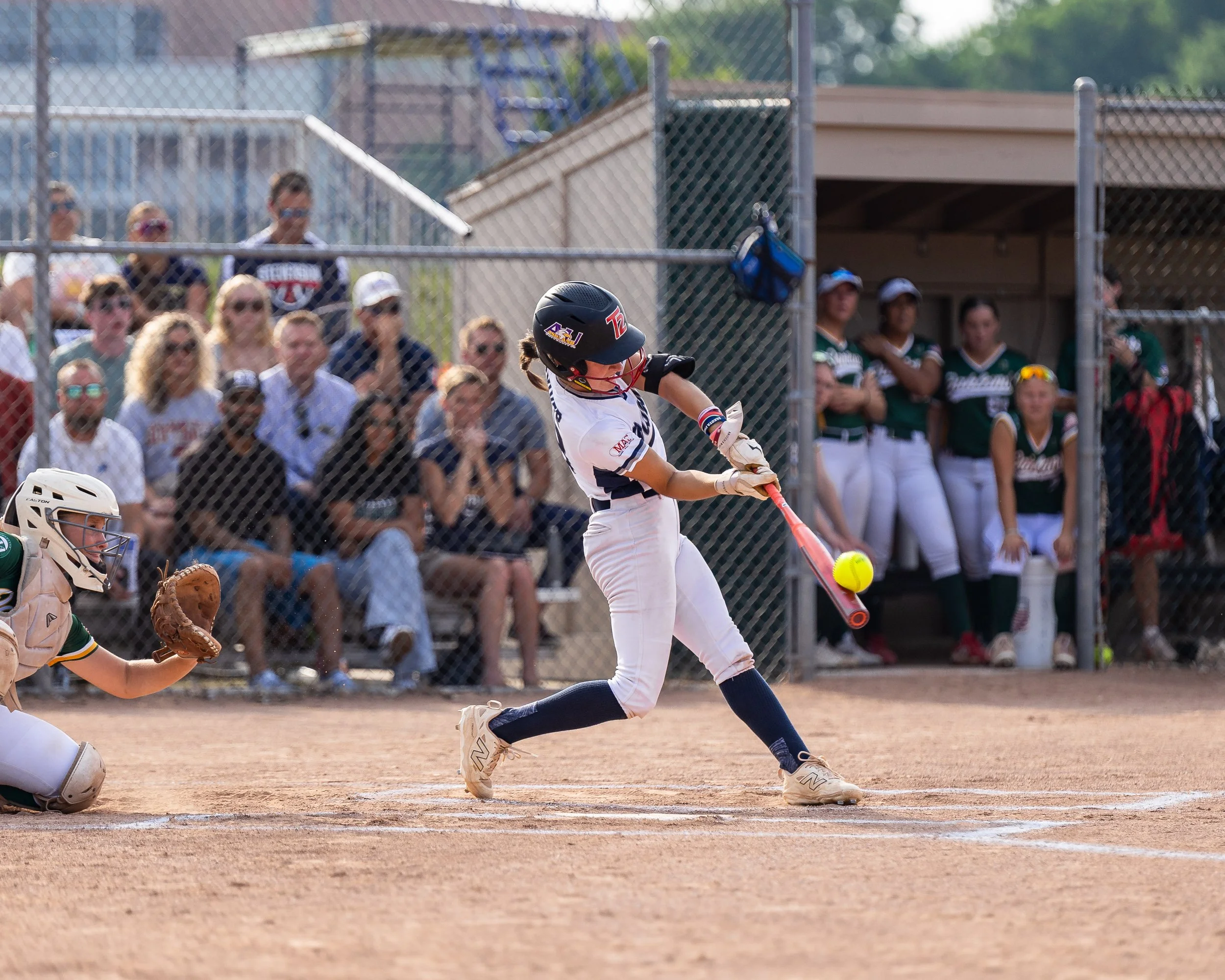 A young girl batting in a softball game, wearing a helmet and uniform, hitting a yellow ball with a pink bat while the catcher is ready behind her. Spectators in sunglasses and team members watch from the dugout and stands.