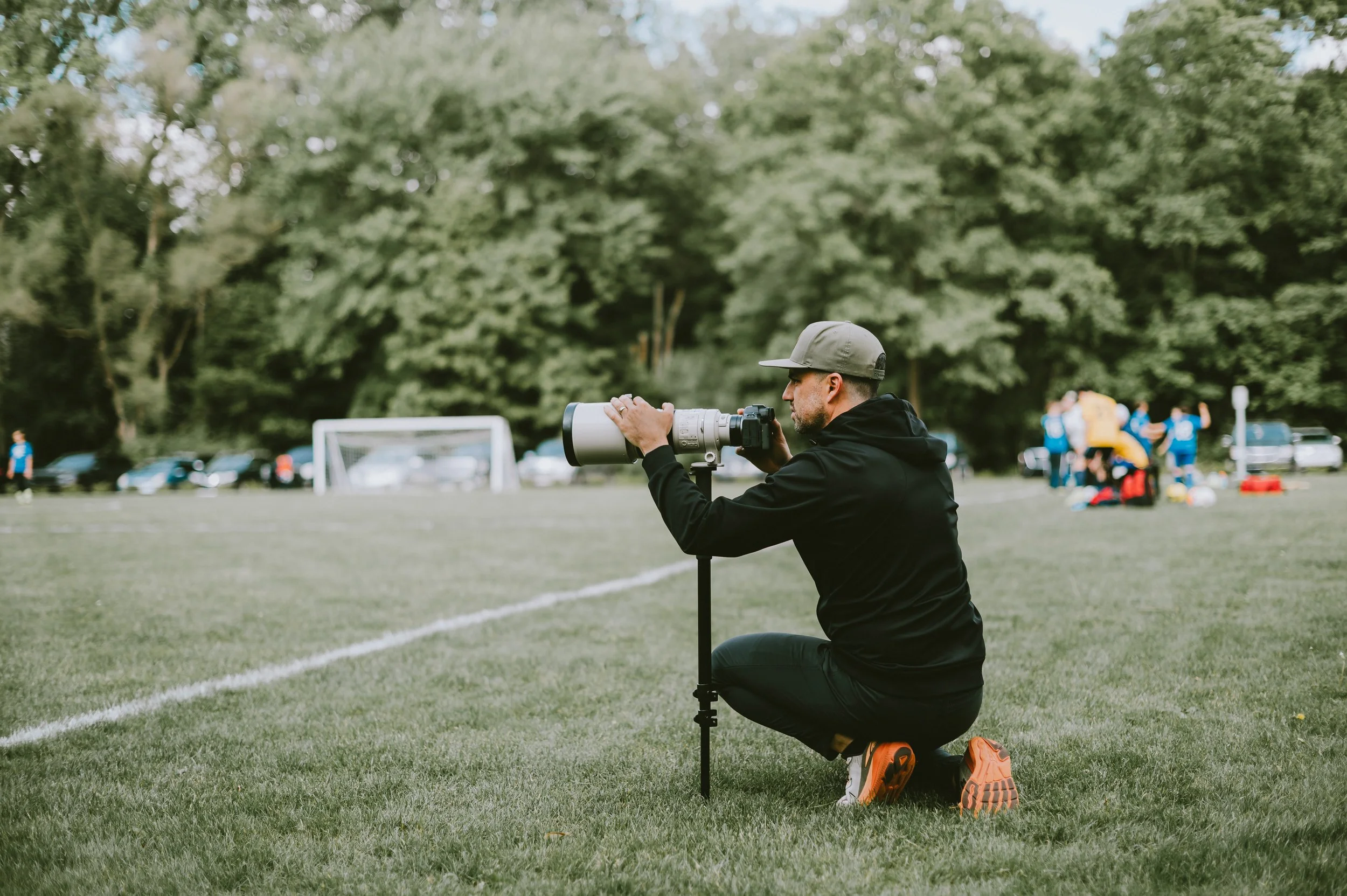 Photographer kneeling on grass with a camera and telephoto lens during outdoor event, with group of people in the background on field.