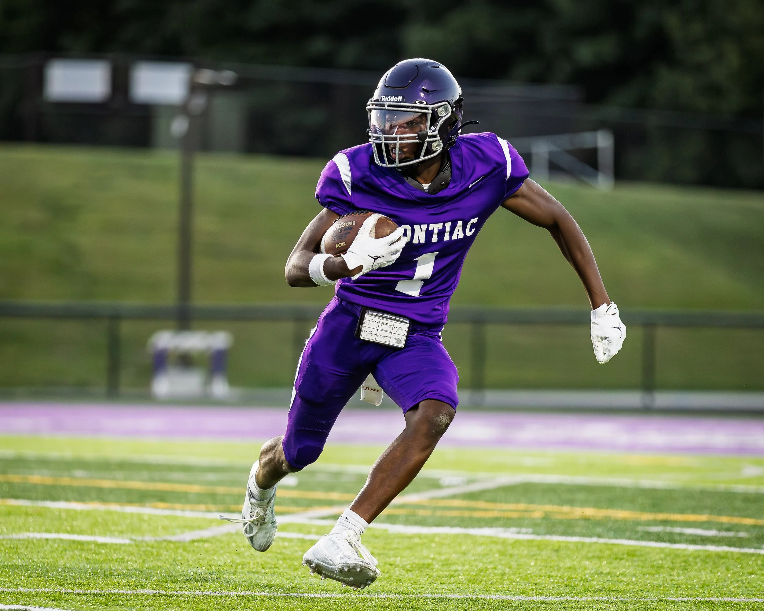 An American football player in a purple uniform with the word 'ONTIAC' on the front is running with a football during a game.