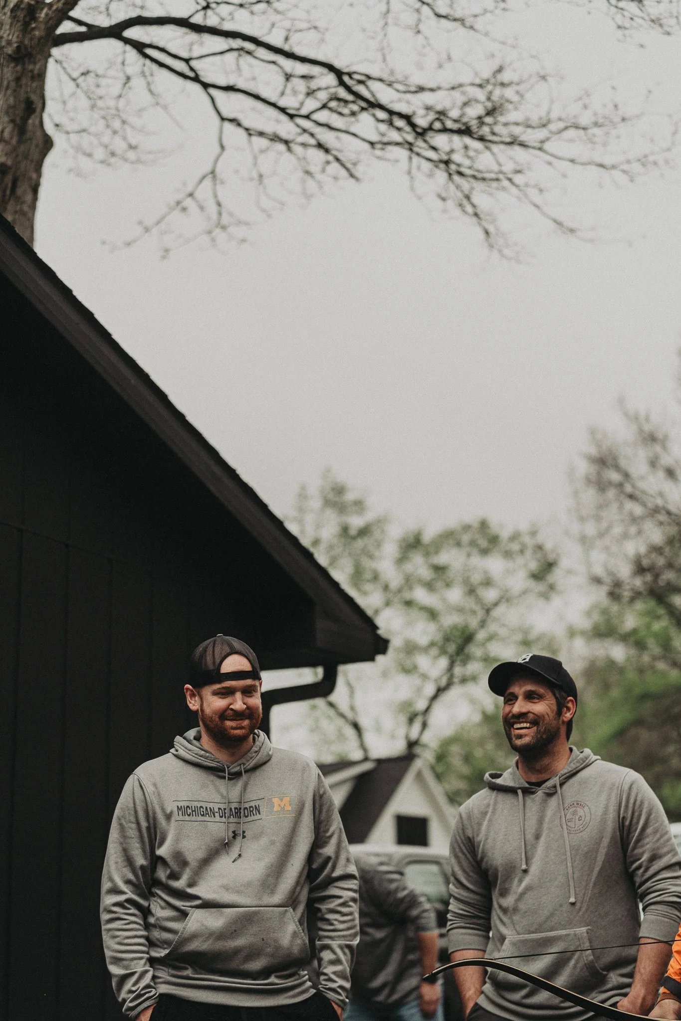 Two men smiling and talking outdoors on a cloudy day, wearing gray hoodies and baseball caps, near a black wooden building with trees in the background.