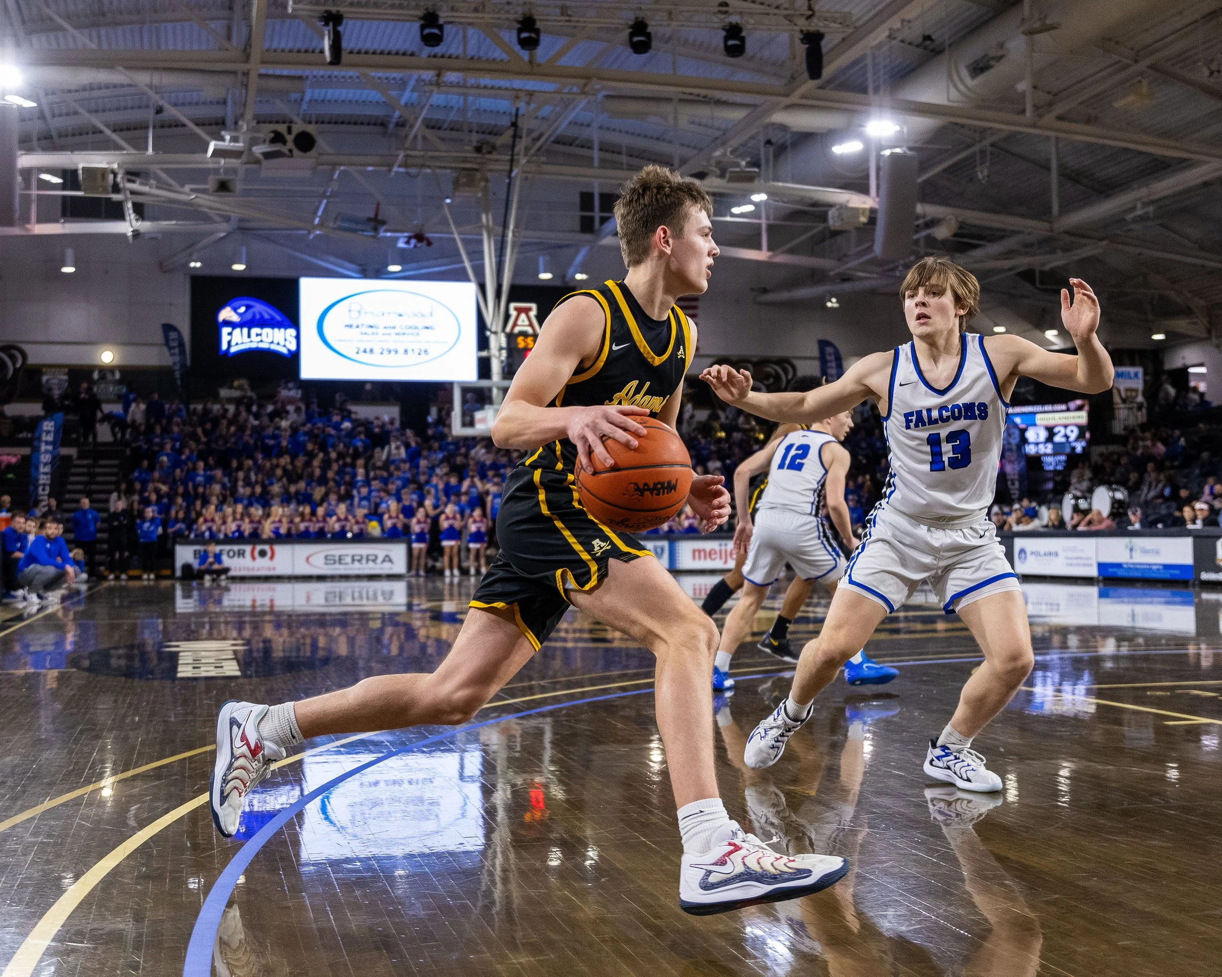 A youth basketball game with players in black and white jerseys on a polished indoor court, with a large crowd and digital scoreboard in the background.