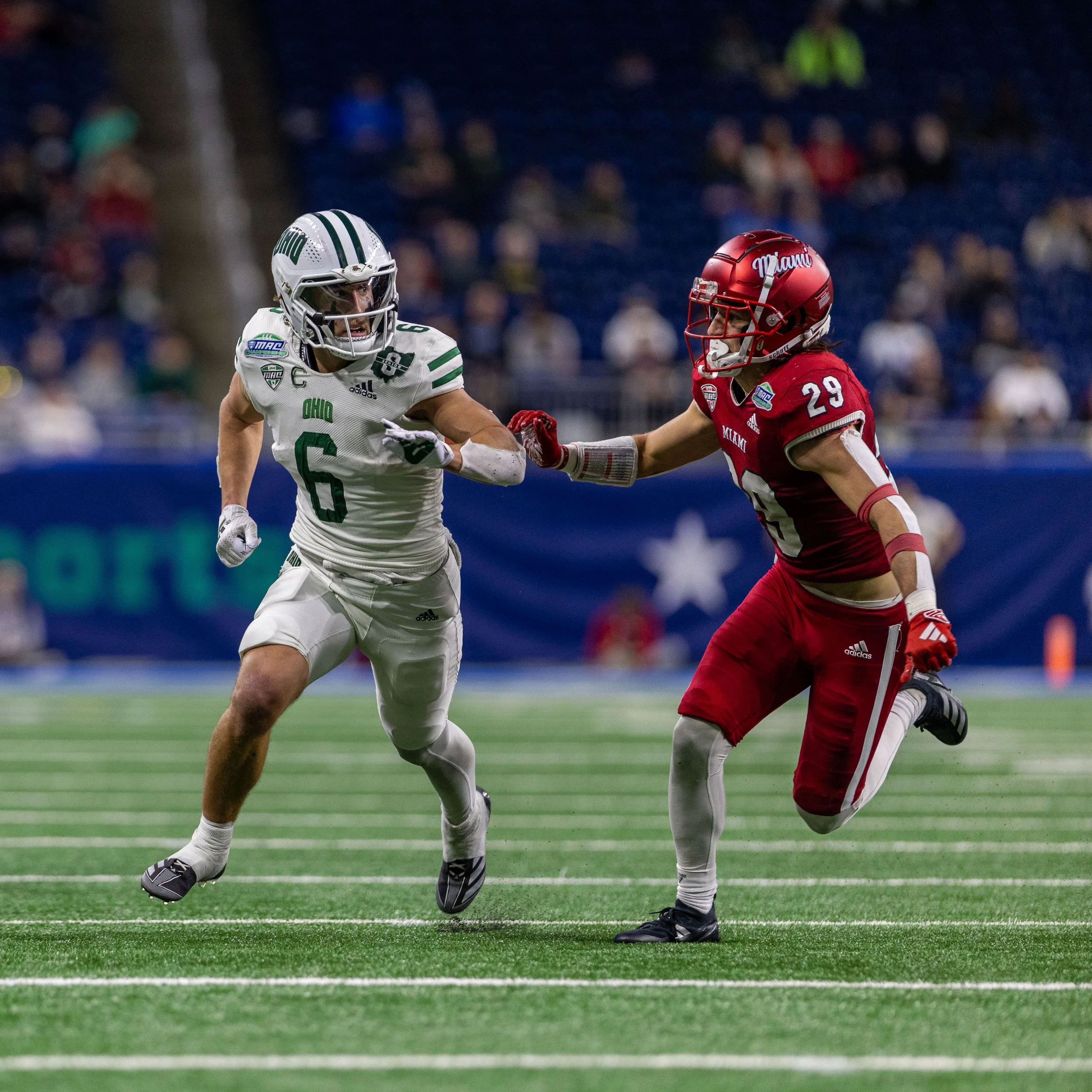 Two football players in action during a game, one in a white Ohio uniform and the other in a red Miami uniform, with a stadium and spectators in the background.