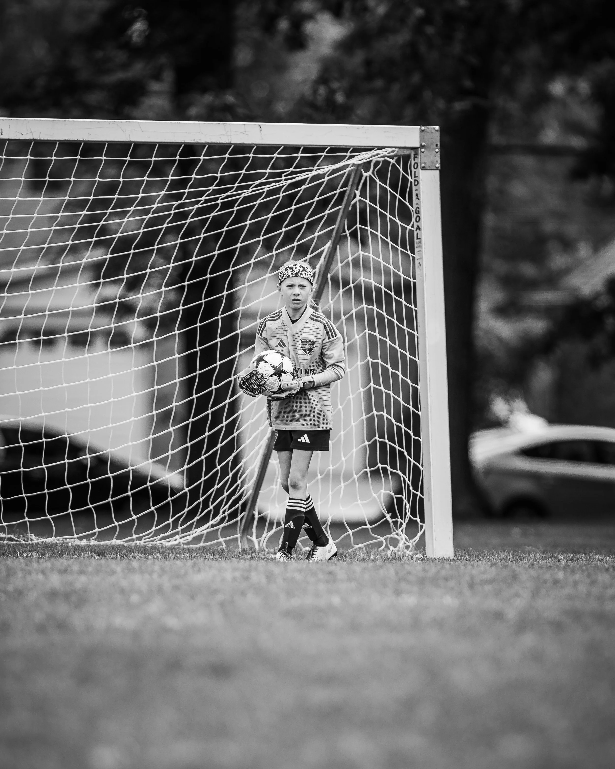A young girl standing in the goal of a soccer field, holding a soccer ball, wearing a goalkeeper uniform and gloves, with a serious expression.